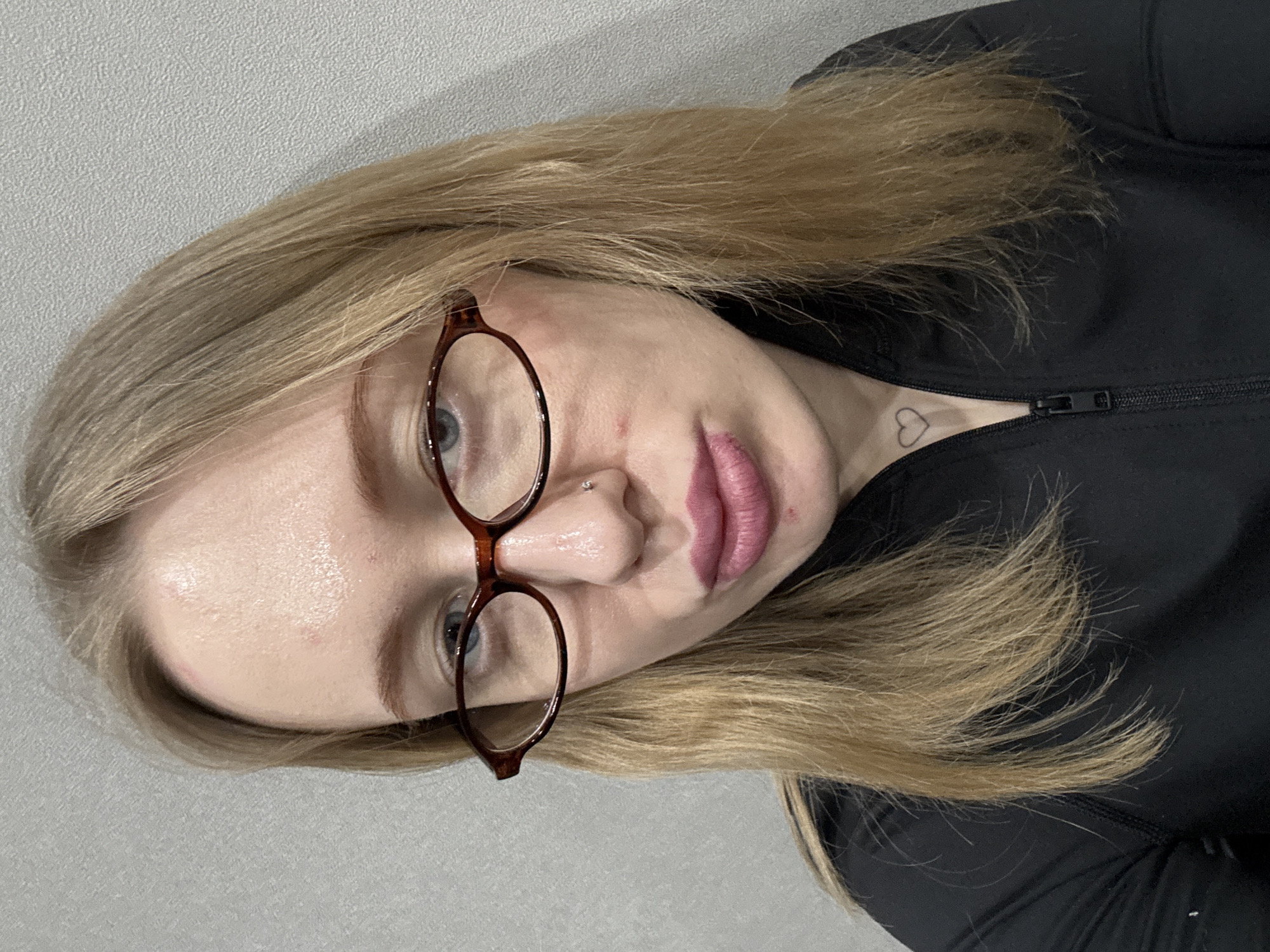 A young woman with blonde hair and blue eyes is wearing brown-rimmed glasses and a black jacket. She has a nose piercing on the left side and a small heart tattoo on her neck. The background appears to be a light grey wall. The location is not identifiable as Kuleshovka, Russia from this image.