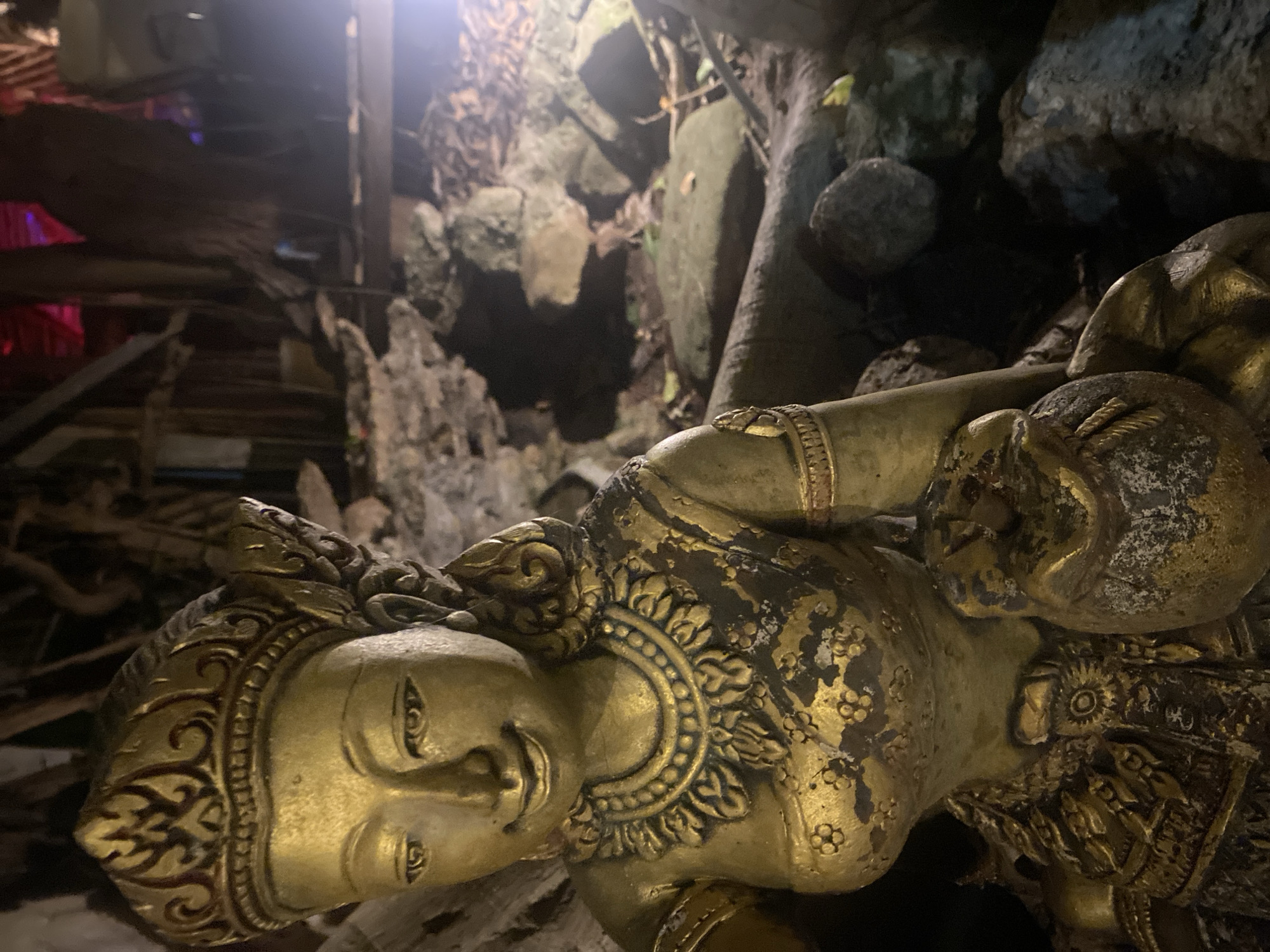A gilded statue of a reclining Buddha is situated within a cave or temple environment in Baan Don Sai, Thailand. The statue shows signs of age and wear, with areas of corrosion and loss of gilding. The surrounding environment is rocky and includes what appears to be tree roots and cave formations. The area is dimly lit, with some lighting coming from above the scene.