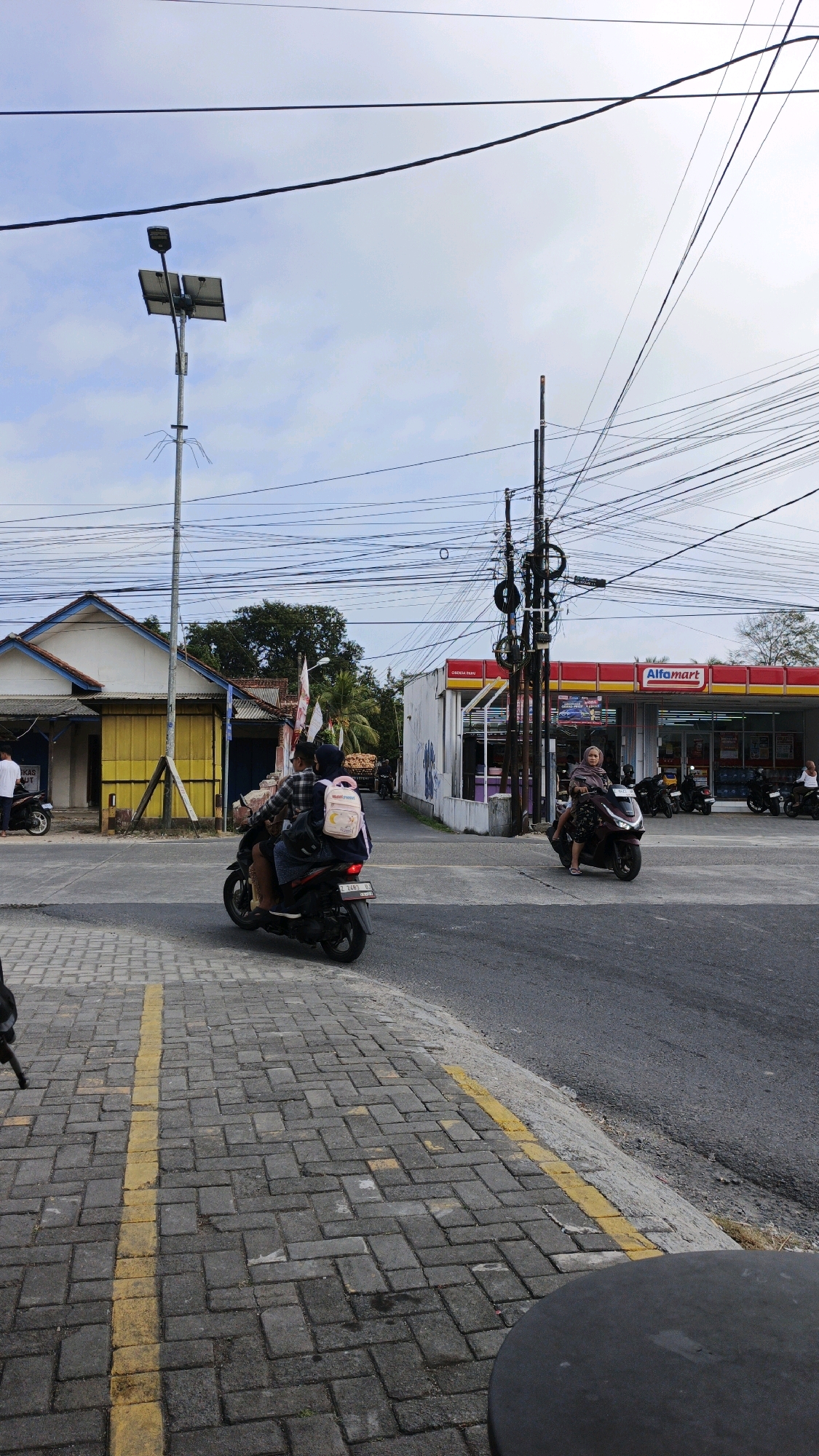 In Cibenda, Indonesia, a street scene unfolds under a cloudy sky. The foreground features a paved sidewalk with a yellow dividing line, while the street is asphalt. Two motorcycles are prominent in the scene. One, a dark scooter, carries two individuals. The person in front wears a plaid shirt, and the passenger behind has a backpack with a cartoon character. This motorcycle is in the left lane, heading away from the camera. Further down the street, another dark scooter is being ridden by a person in a headscarf, heading towards the camera.
To the right, an Alfamart convenience store is visible with its red and yellow signage. Several motorcycles are parked in front of the store. On the left side of the street, a building with white and yellow facades stands, with a motorcycle parked in front of it. A solar-powered street lamp with two panels is positioned near the left side of the frame, and numerous electrical wires crisscross the sky, supported by utility poles. The overall environment suggests a commercial or residential area in an Indonesian town.