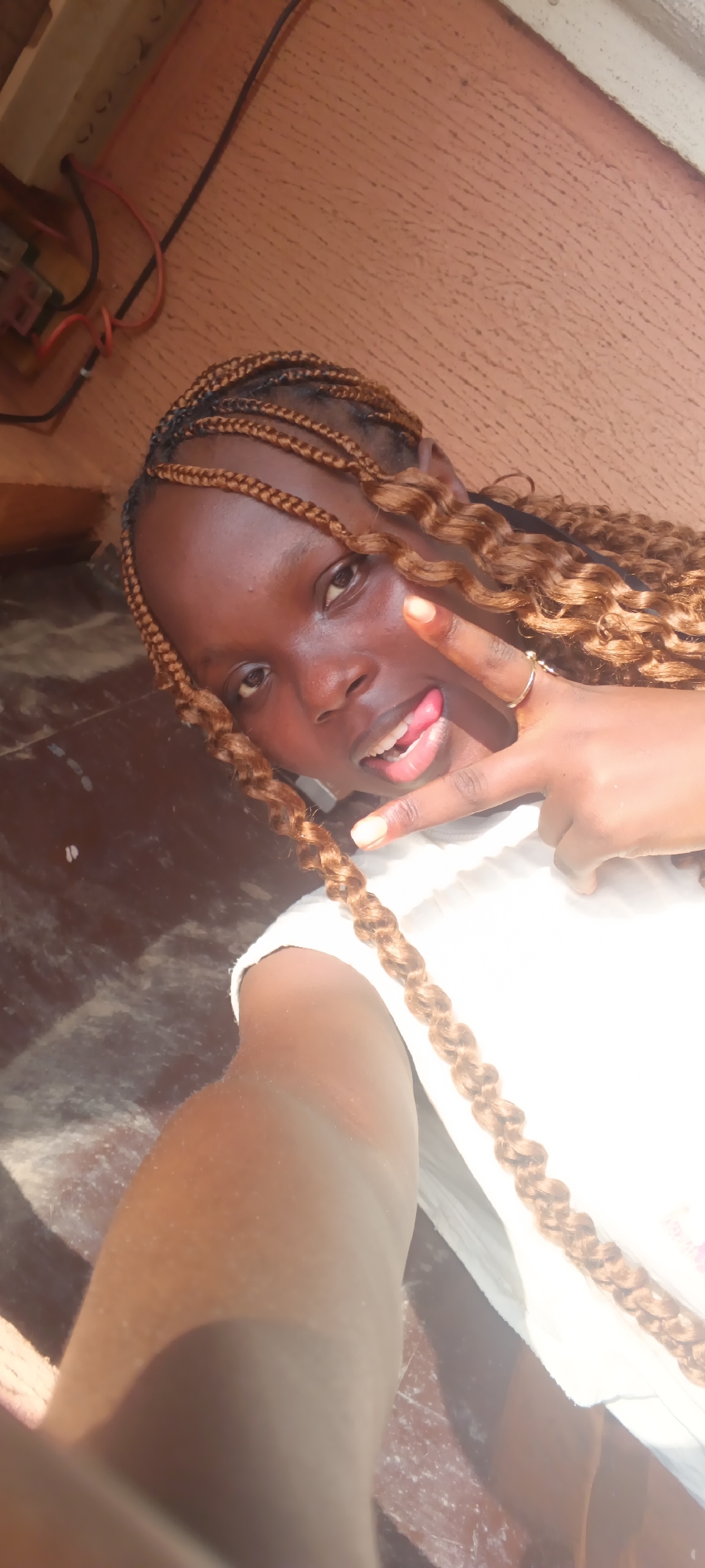 This is a close-up selfie of a young person with dark skin and light brown braided hair, wearing a white top. She is smiling playfully, with her tongue slightly out, and makes a peace sign with her left hand; a ring is visible on her ring finger. Her braided hair features small, neat cornrows near the scalp and thicker, wavy or curly braids that frame her face and fall over her shoulder.
The strong, bright lighting indicates it is daytime, casting highlights on her skin and clothing. The background consists of a textured, orangey-brown wall or surface in the upper portion. On the far left, black and red electrical wires are visible, connected to what appears to be a power strip or an electrical component. The lower part of the background is a darker, speckled surface, possibly a floor or table. The overall impression is one of cheerfulness and casualness. No text is visible in the image.