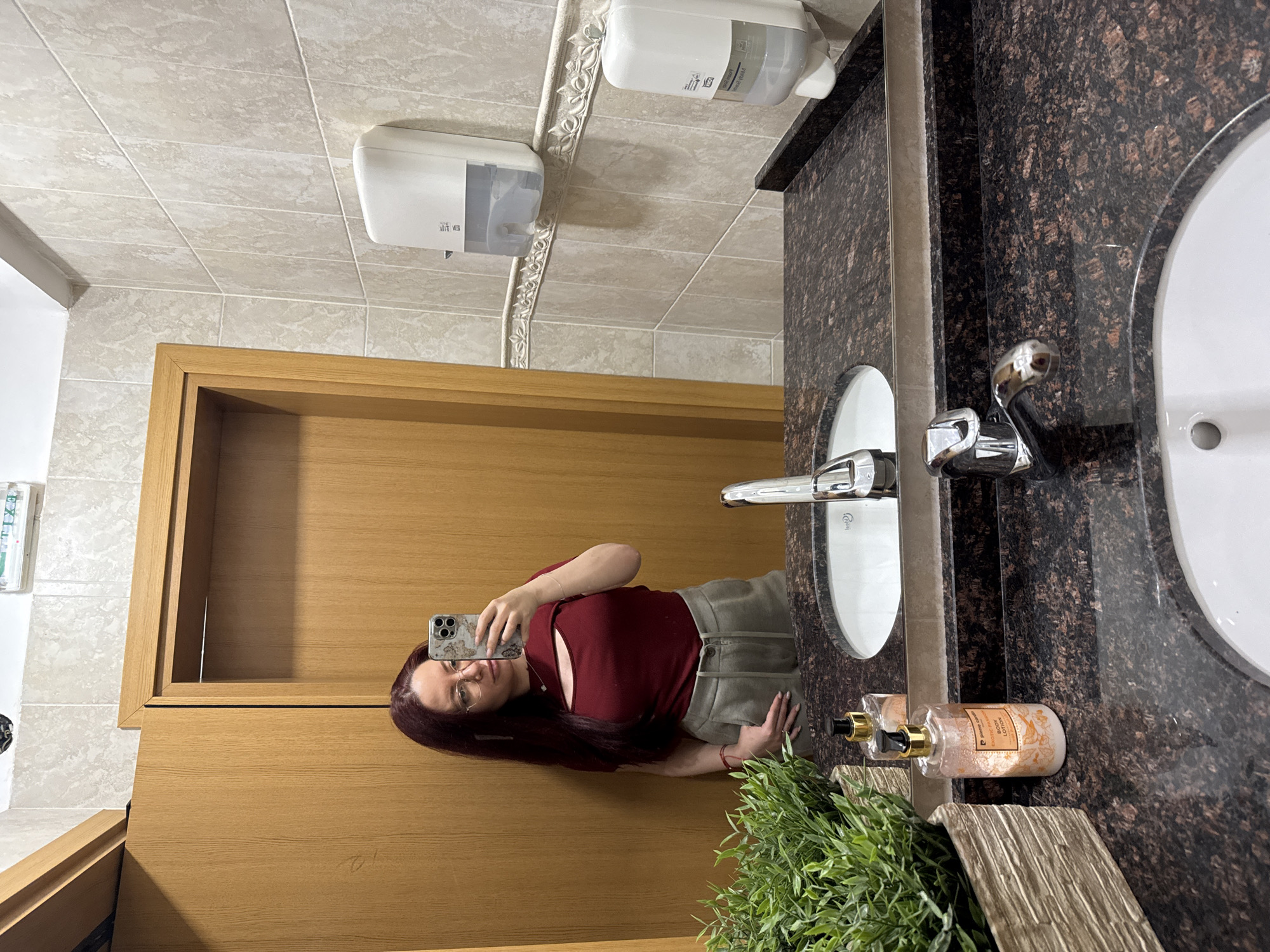 A woman is taking a selfie in a bathroom mirror. She is wearing a dark red, short-sleeved top and khaki pants. The bathroom has tiled walls, a wood-paneled door, a dark speckled countertop with a double sink, a faux plant arrangement, and toiletries on the counter. There are two soap dispensers on the wall. There is no definitive evidence in the photo to suggest that this bathroom is in Sofia, Bulgaria.
