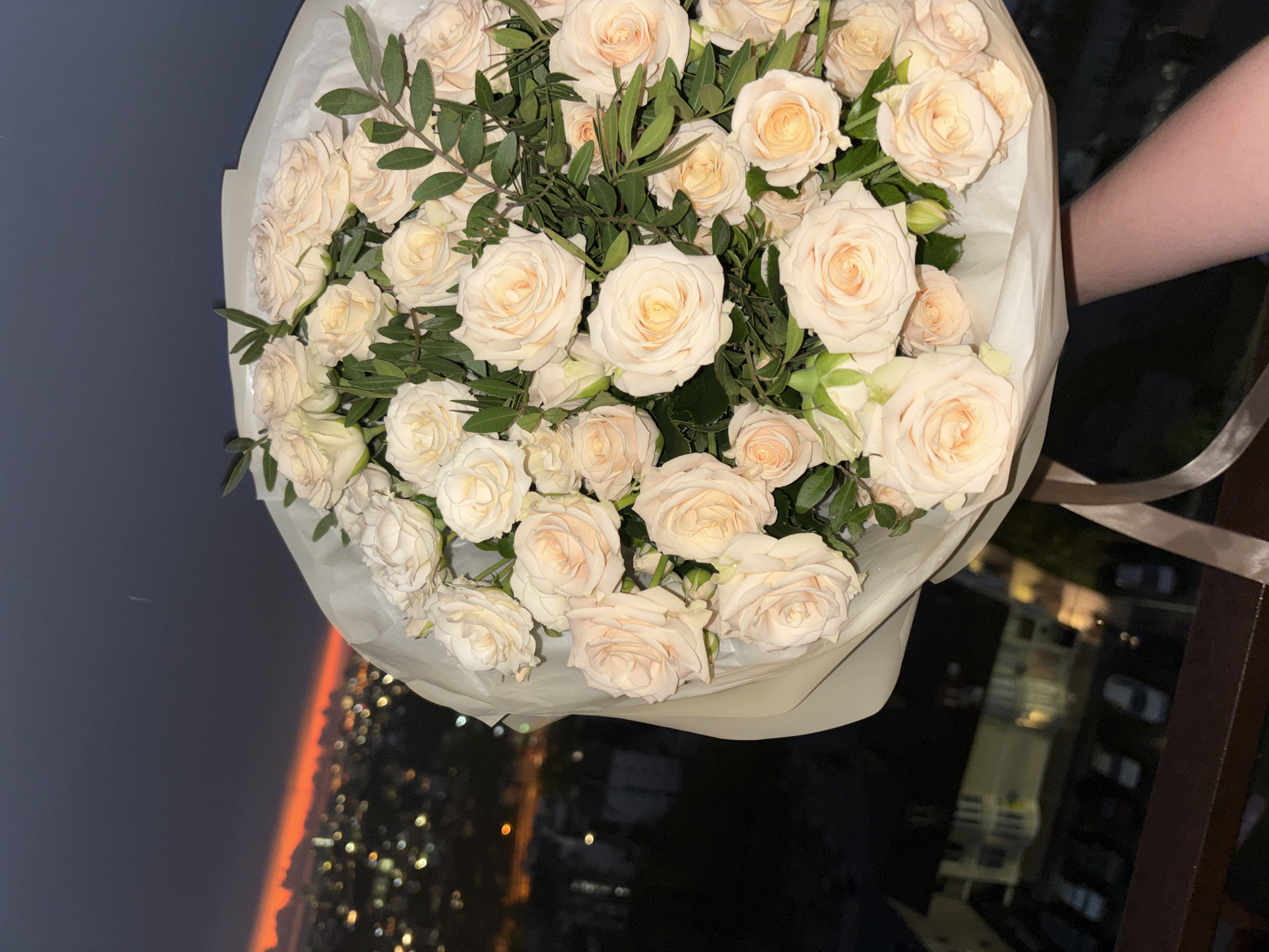 A bouquet of white roses is held against the backdrop of a nighttime urban landscape. The roses, wrapped in light-colored paper, are the main subject, with visible green foliage interspersed among the blooms. The urban background appears to be a city at night, with scattered bright lights suggesting buildings and streets. An orange-tinted horizon is faintly visible in the distance, possibly indicating sunset or sunrise. The image is taken from an elevated position, suggesting a balcony or high floor. There is a glimpse of a light-skinned human arm holding the bouquet. The specific location as Sochi, Russia, is not evident from the image itself.