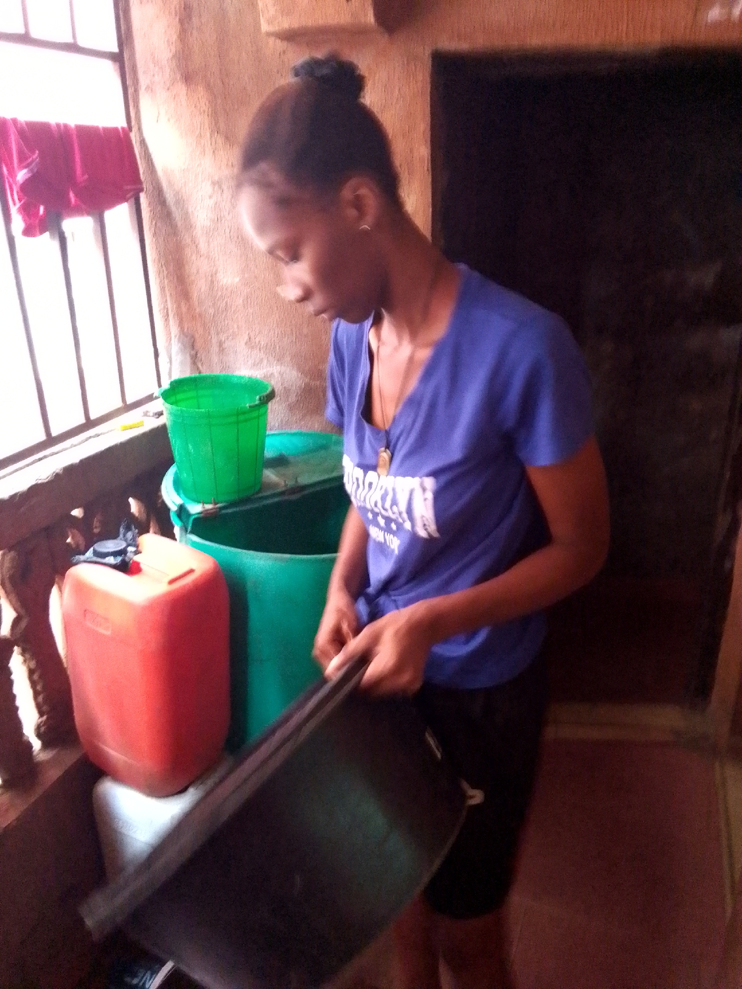 A person stands indoors next to a balcony, attending to items including green buckets, a red jerrycan, and a dark-colored tray. The person, likely female, wears a blue t-shirt and black shorts. A window with a red garment hanging is visible. A dark doorway is to the right of the person.