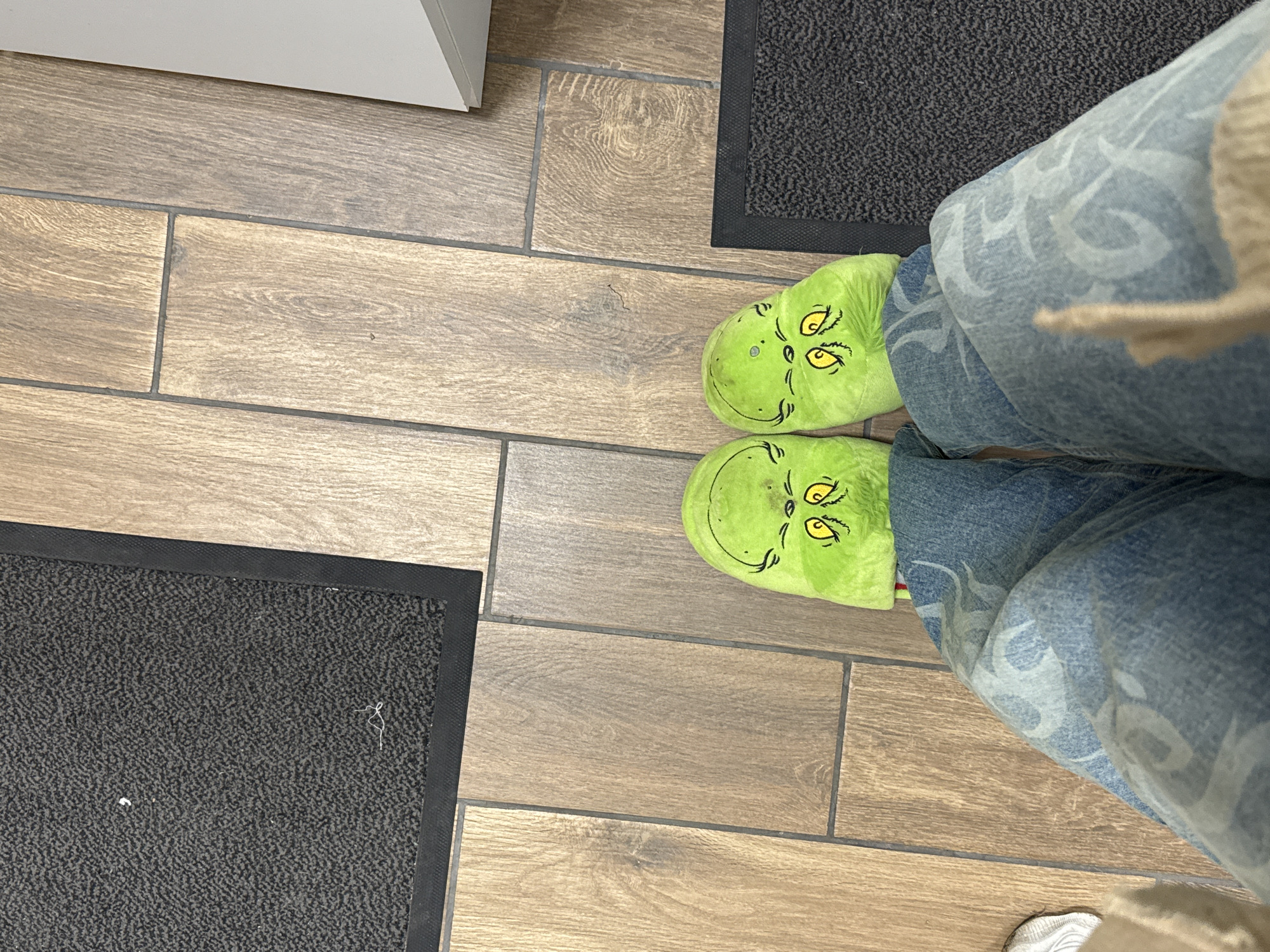 The image shows a person wearing light blue jeans with a white design on the right leg and green Grinch slippers. The person is standing on a light brown tile floor with dark grey grout lines. There are two black mats with a textured surface visible on either side of the slippers. A white cabinet is partially visible in the upper left corner. The location is not possible to determine from the provided image.