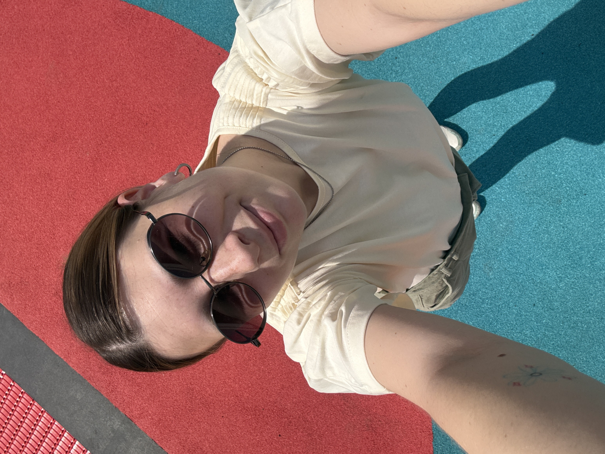A person takes a selfie outdoors. The person is wearing sunglasses and a light-colored shirt. A blue and red surface is visible behind them. A hand shadow is visible on the blue surface. The surface appears to be made of a rubbery material. The photo appears to have been taken at Gorky Park in Moscow, Russia, given the characteristic red and blue ground covering.