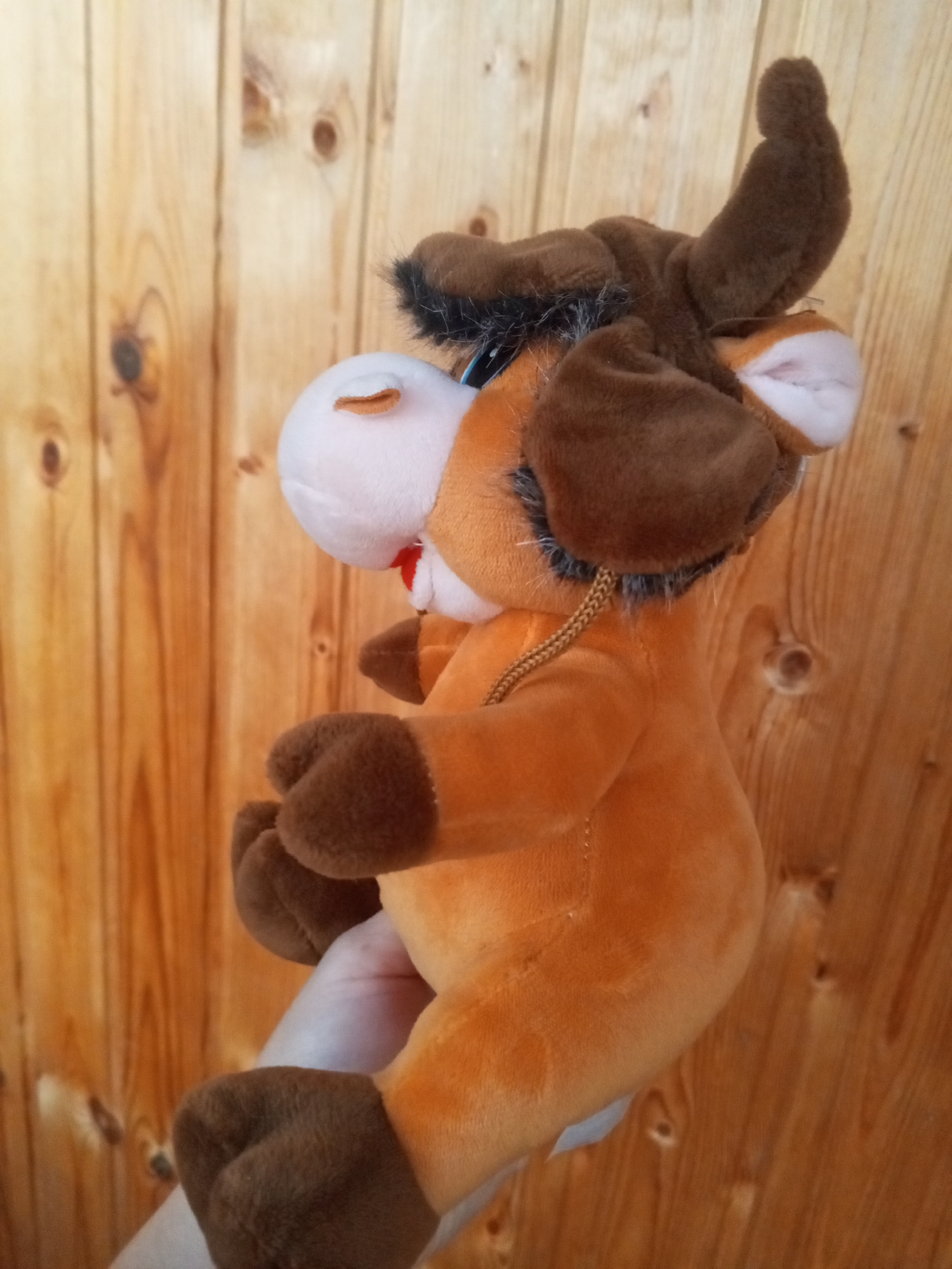 A plush toy bull is being held by a person's hand against a wooden background. The bull is brown and orange, with white and pink accents on its face. It has closed eyes and a small red mouth. The background is made of vertical wooden planks with visible knots. The image appears to be taken indoors.