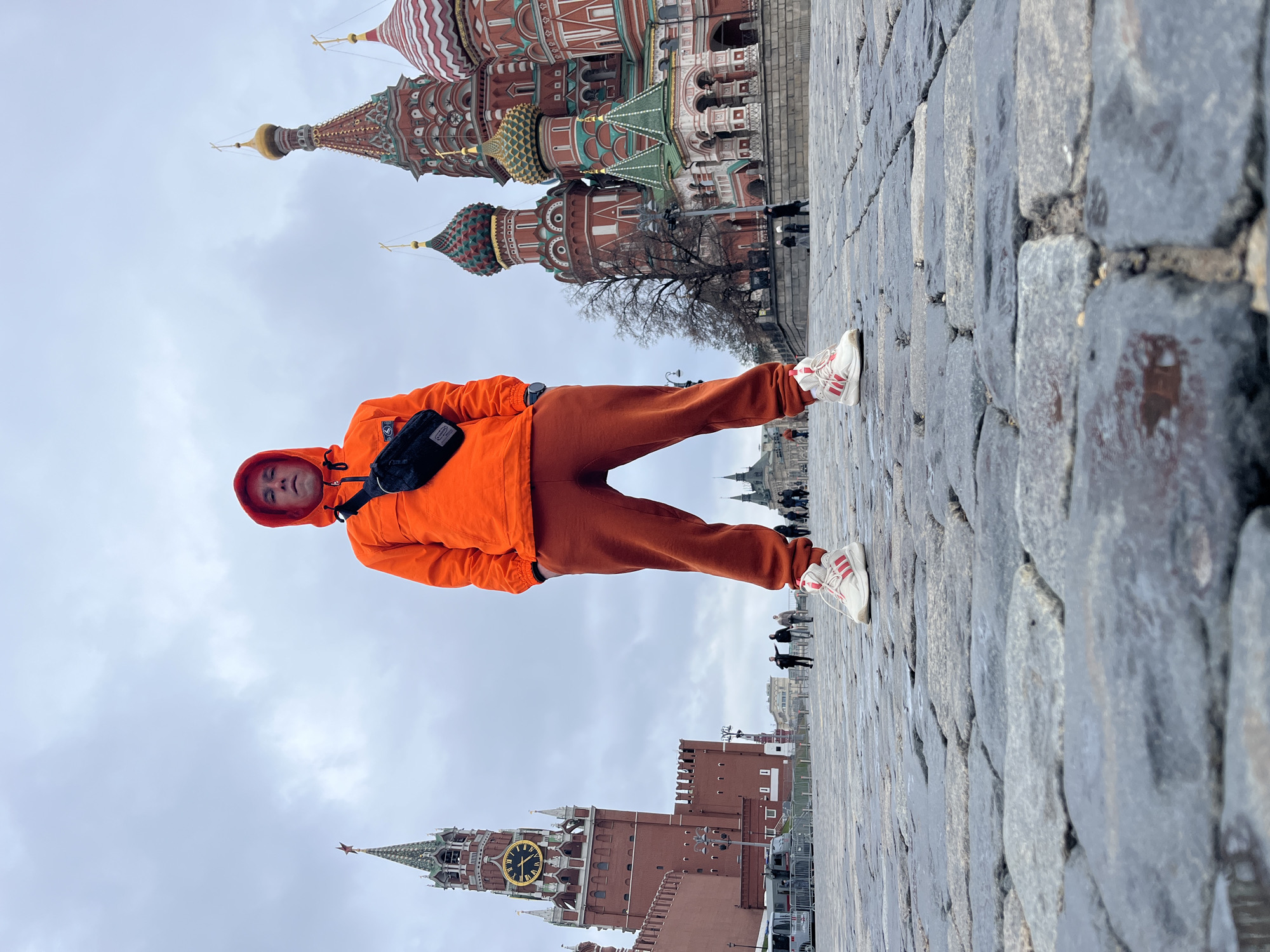 A man in an orange hooded jacket and orange pants poses in front of St. Basil's Cathedral and the Kremlin Clock Tower in Moscow, Russia. The man is wearing white sneakers with red stripes and a black shoulder bag. He stands on a cobblestone surface. Several people are walking in the background. The sky is overcast.