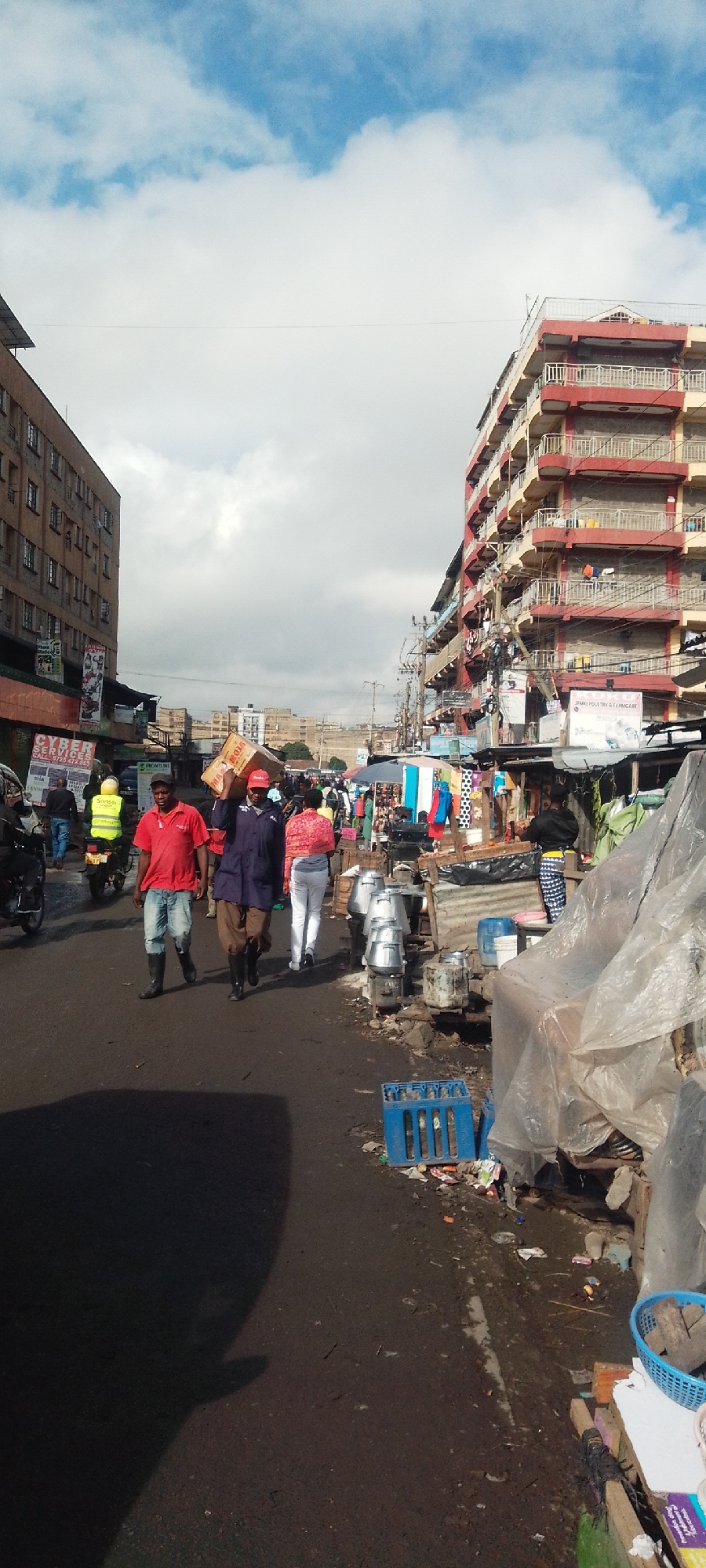 This image depicts a bustling urban street scene under a partially cloudy sky with patches of blue. The location is an active commercial or market district within Nairobi, Kenya, evidenced by the multi-story buildings and numerous street vendors.
On the left, a multi-story building with a brownish-grey facade and multiple windows features signage on its lower level, including a partial "CYBER SERVICES" sign. On the right, a prominent multi-story building displays a red and pale yellow exterior with multiple balconies, some adorned with hanging laundry. A sign for "JENKI POULTRY & PHARMACARE" is visible on the ground level of this building. Numerous tangled overhead utility lines crisscross between the structures.
The street itself is an asphalt surface, appearing worn and dirty with scattered debris, including plastic items and refuse, particularly along the right side near the market stalls. A large, dark shadow dominates the lower left quadrant of the road. A blue plastic crate is visible on the ground on the right.
Approximately ten to fifteen individuals are visible. In the midground, a male in a red t-shirt, light shorts, and black boots walks towards the viewer on the left side of the road. Alongside him, another male, wearing a dark blue long-sleeved top, light brown trousers, a red cap, and black boots, carries a large cardboard box on his right shoulder while walking towards the viewer. Behind them, a female in a patterned top and white trousers walks away. Further back on the left, a person wearing a bright yellow reflective vest is operating a motorcycle, moving away from the viewer.
The right side of the street is lined with informal market stalls, constructed from makeshift materials, some covered with tarpaulins or clear plastic sheeting. One prominent clear plastic sheet covers a stall in the foreground. Several large, silver-colored metal containers are stacked on crude supports near the foreground market area. Clothing items, including colorful fabrics, are displayed under an umbrella in the midground stalls. A vendor in a black top and patterned skirt attends a stall further back on the right.