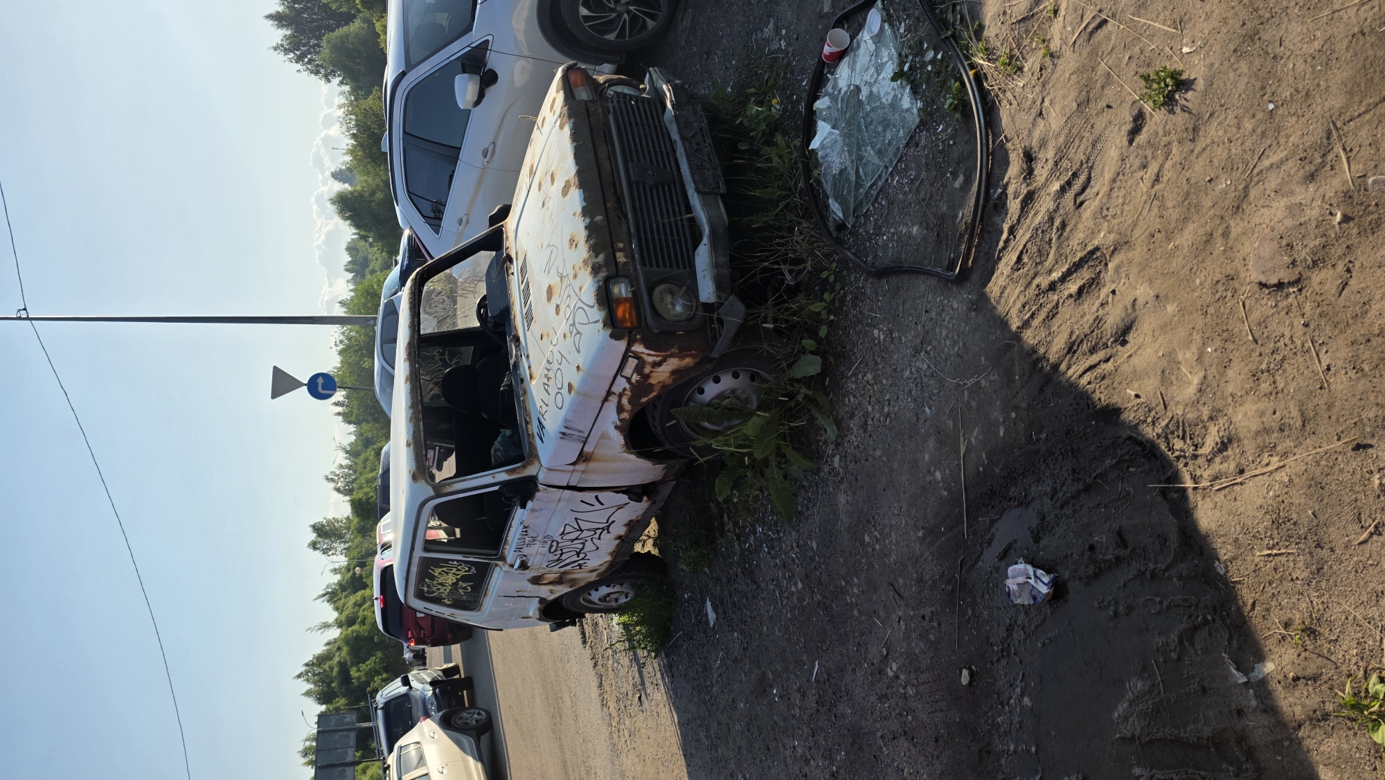 An abandoned, rusty, white Lada Niva is parked on a dirt and gravel surface next to a paved road with other vehicles in the background. The car's windshield is missing and shattered glass lies on the ground in front of it. The car has graffiti spray-painted on its exterior. There is vegetation growing around the car and on the roadside. The sky is clear and blue with a few white clouds. The scene appears to be in a rural or suburban area.