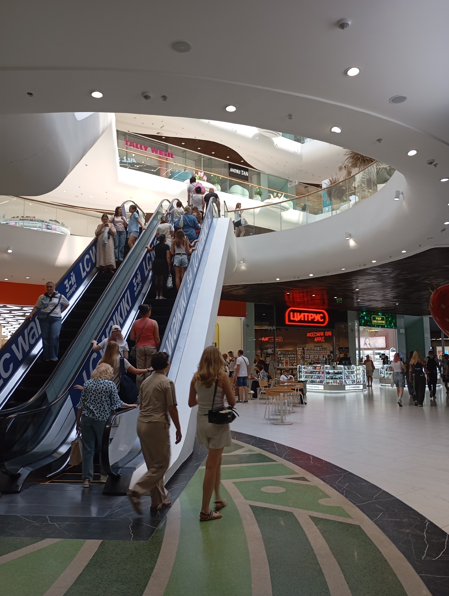 The image depicts the interior of a modern shopping mall. In the foreground, a group of people are ascending an escalator. The escalator steps are emblazoned with the text "LC Waikiki". To the left of the escalator, another escalator is visible with similar branding and people ascending. A woman in a blue and white patterned shirt and jeans is standing on the stationary part of the escalator. Further up, several individuals are ascending the escalator, including a woman in a light brown jumpsuit.
In the mid-ground, beyond the escalators, several shops are visible. One prominent shop features a large red neon sign with the Cyrillic text "ЦИТРУС", indicating a store selling electronics and accessories for Apple products. Adjacent to it, another shop displays a green sign with Cyrillic text that appears to say "ТКР ЗОЛОТО" and an advertisement for a jewelry store. People are walking through the mall area, and seating areas with tables and chairs are present.
The architecture of the mall is characterized by curved white ceilings and modern lighting. The floor is a mix of dark marbled tiles and a patterned green terrazzo. The overall ambiance is bright and busy, suggesting a typical shopping day. This scene is consistent with the interior of the Nikolsky Shopping Centre in Kharkiv, Ukraine, a well-known retail complex.