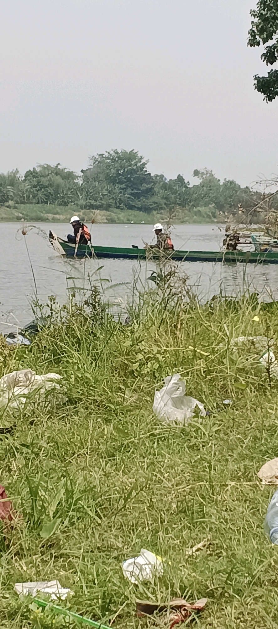In Laren, Indonesia, a serene river flows under a hazy sky. The riverbank is overgrown with lush green grass, littered with white plastic bags and debris. Two individuals in a narrow green boat navigate the calm waters. Both wear white hats and orange life vests, suggesting safety precautions. The person in the front is actively engaged in fishing, casting a net into the water. The other individual sits further back, observing. The backdrop features a dense line of trees and foliage, indicative of a natural, rural landscape. The overall scene depicts a peaceful day of fishing, juxtaposed with the unfortunate presence of human-generated waste along the riverbank.