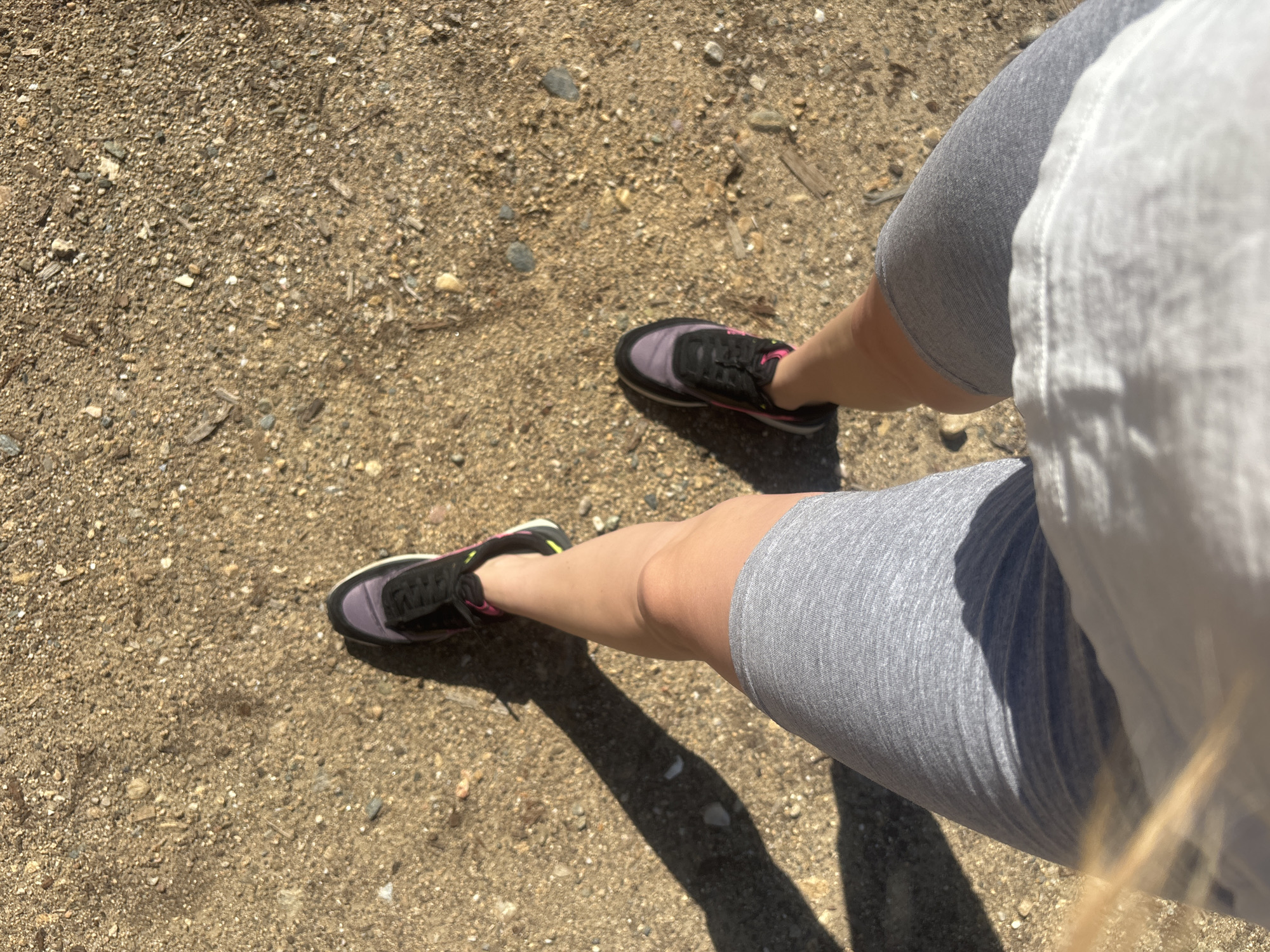 The image shows a first-person perspective of a person walking on a sandy and gravelly path in Mikro Doukato, Greece. The person is wearing grey athletic shorts, a white t-shirt (partially visible), and athletic shoes with purple, black, and pink accents. The sun casts a long shadow to the left of the person's legs, indicating it is likely daytime. The environment is outdoors, characterized by loose soil and small stones.