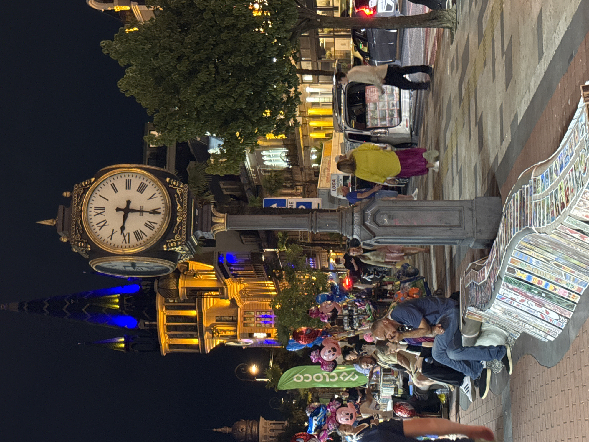 The image depicts a street scene at night in Batumi, Georgia, likely on Batumi Boulevard, a prominent tourist area. A large, ornate clock tower with illuminated blue and yellow lights stands prominently in the background, an iconic landmark of the city. In the foreground, a decorative bench, adorned with numerous colorful tiles depicting various scenes and images, curves along the walkway.
Several people are visible. To the left, a vendor is selling balloons, including Mickey Mouse and Spider-Man themed ones, to a group of people. Some individuals are seated on the bench, while others are standing or walking along the sidewalk. A woman in a bright yellow dress and a purple skirt walks past a van with its rear hatch open, which appears to be a mobile vendor or display. Several cars are parked or driving on the street. The overall atmosphere suggests a lively evening gathering, possibly during a tourist season. The street is paved with brick and has a patterned sidewalk. Trees line the street, providing some shade. The scene is well-lit by streetlights and the illumination from the buildings.
