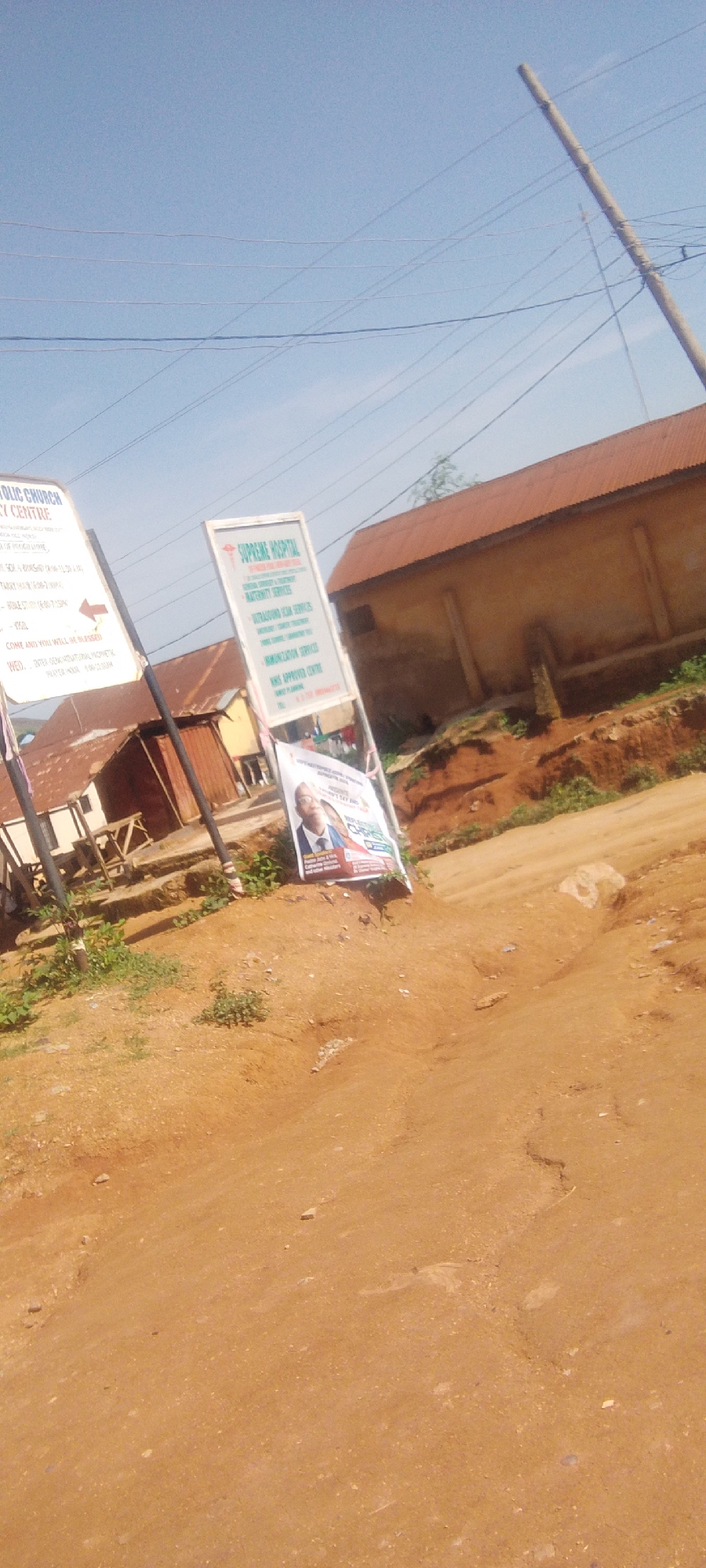 In Kwamba, Nigeria, on a dirt road under a clear blue sky, a scene unfolds featuring two signs and a building. To the left, a white sign with black text reads "CATHOLIC CHURCH PRAYER CENTRE," listing various activities and prayer times. A red arrow points to the right. Beside it, a taller, white sign advertises "SUPREME HOSPITAL," detailing services like general surgery, maternity, and ultrasound. Below this, a banner features two men in suits, one with a microphone, and text that is partially obscured but includes the name "CHRIS." The building in the background is a single-story structure with a rust-colored corrugated metal roof and light-colored walls, situated on a slight incline. The surrounding environment is characterized by dry, reddish-brown earth, sparse green vegetation, and overhead power lines supported by a utility pole. The overall impression is of a semi-rural or developing urban area.