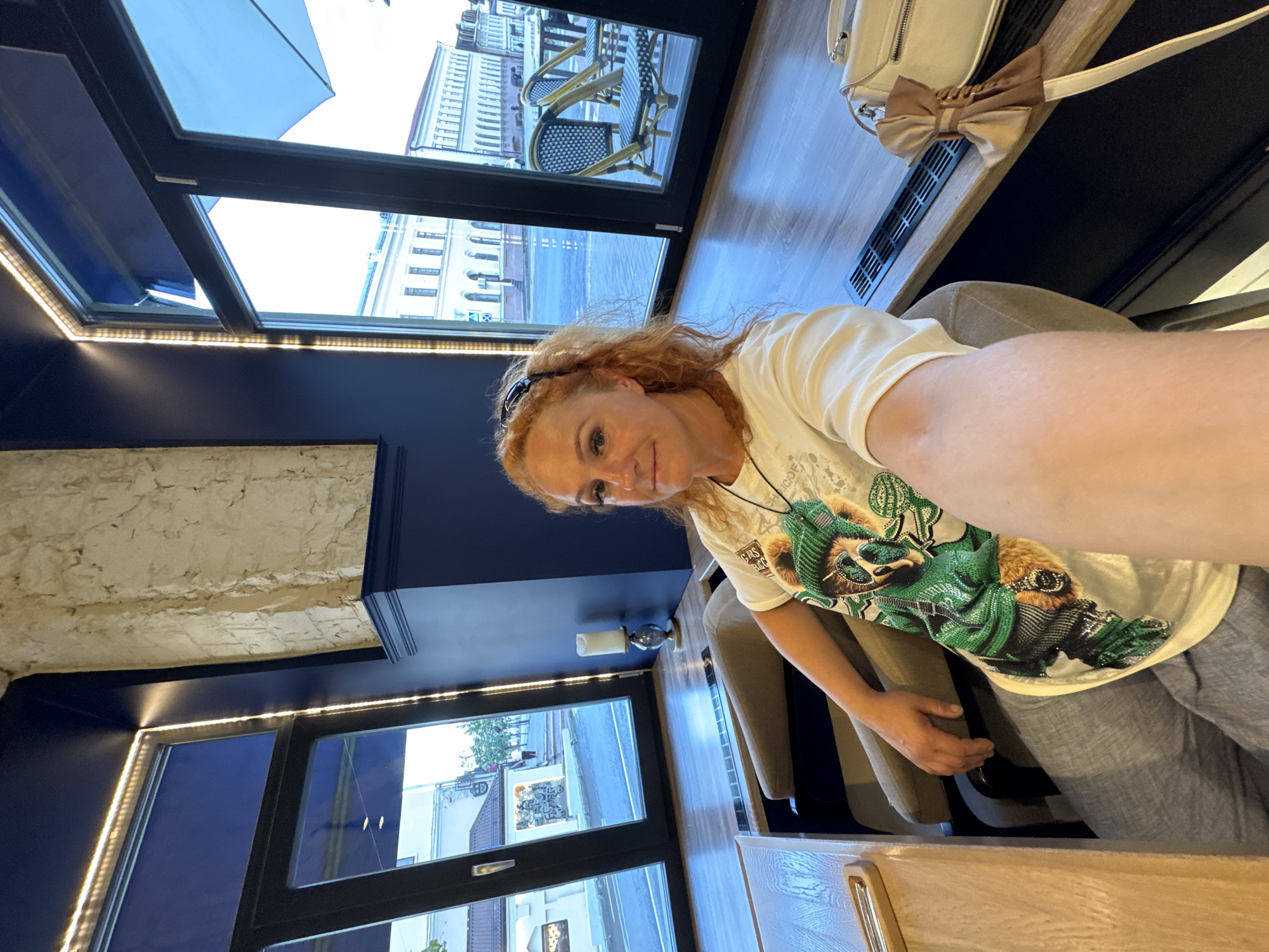 A woman with red, curly hair is taking a selfie in what appears to be a cafe or restaurant in Kazan, Russia. She is seated in a comfortable chair, wearing a light-colored t-shirt with a graphic print of a bear. The cafe has large windows that look out onto a street with several buildings. The interior is modern, with blue walls and wooden furniture. There is a decorative candle on a stand near the window, and a white handbag with a bow is placed on the table beside her. The lighting in the cafe is a mix of natural light from the windows and artificial light from LEDs installed along the ceiling.