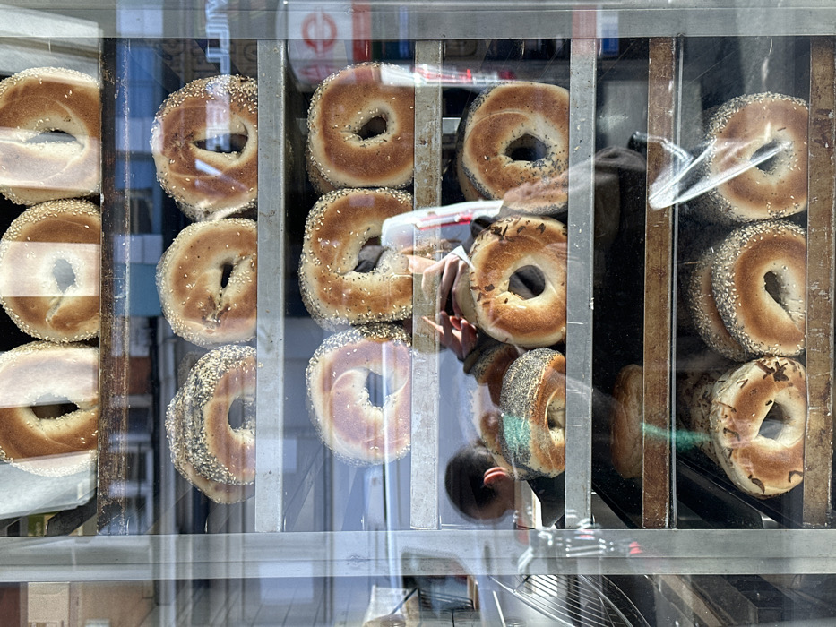 A shop window features a multi-tiered metal display case stocked with numerous freshly baked bagels. Bagels with various toppings, including sesame seeds, poppy seeds, and plain varieties, are arranged on at least five shelves, some stacked. Reflections on the glass surface show the hands and camera of a person capturing the image, along with a partial view of a street scene outside, including architectural elements and a faint vehicle outline on the left. Another individual's head is faintly reflected on a lower shelf. A distinct red circular Transport for London (TfL) roundel logo is clearly reflected in the upper right portion of the glass, identifying the location as the city of London, United Kingdom.