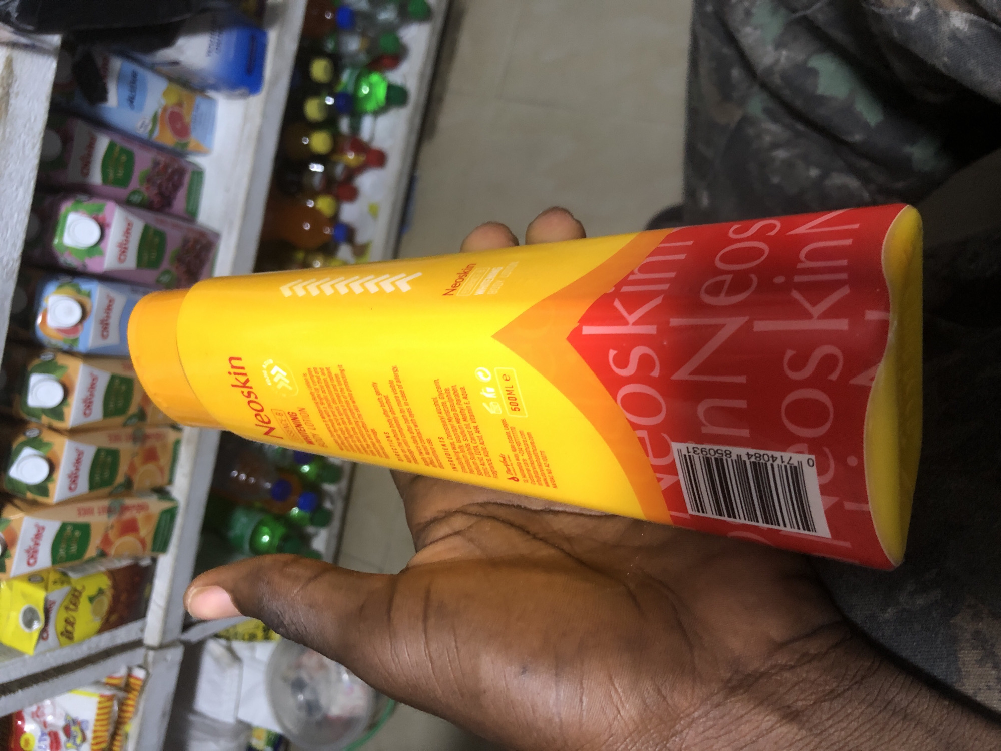 In Ado, Nigeria, a person's hand is holding a bright yellow bottle of Neoskin body lotion. The bottle has a red section with the brand name repeated in a lighter red. The product's label contains information about ingredients and directions for use, and a barcode is visible on the lower part of the bottle. In the blurred background, shelves are stocked with various bottled drinks and cartons, suggesting a retail environment like a convenience store or market.