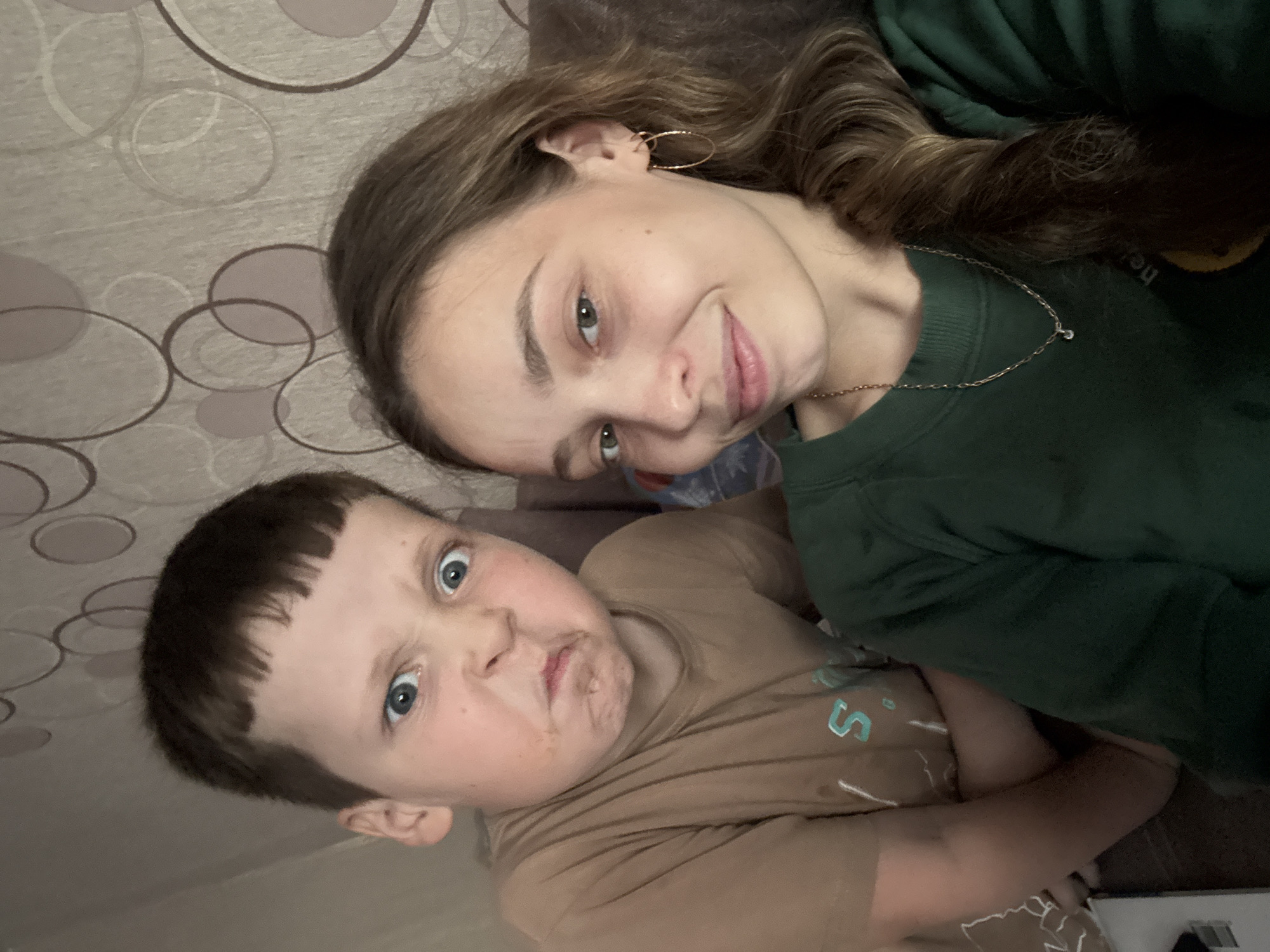The image is a selfie taken indoors, featuring two individuals. On the left is a young boy with brown hair and a tan t-shirt, making a humorous, grumpy face with his eyes wide open and mouth contorted. His expression suggests playfulness. To the right is a young woman with long, brown hair styled in a braid, wearing a dark green sweatshirt and a gold necklace. She is smiling gently at the camera. Behind them, the background consists of a wall with a patterned wallpaper featuring abstract circles. The lighting is dim and appears to be artificial. There is no indication of the specific location within the city of Kemul, Russia.