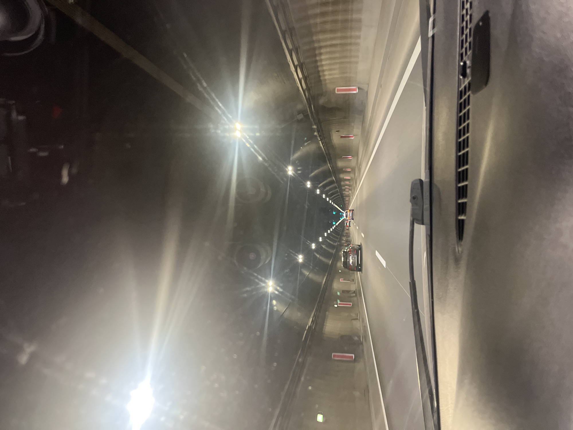 The image depicts a tunnel, possibly underground, illuminated by rows of lights running along its length. Several cars are visible in the distance. The viewpoint is from inside a vehicle or structure, with parts of it visible on the right side of the frame. The scene is suggestive of an urban or transportation infrastructure environment.