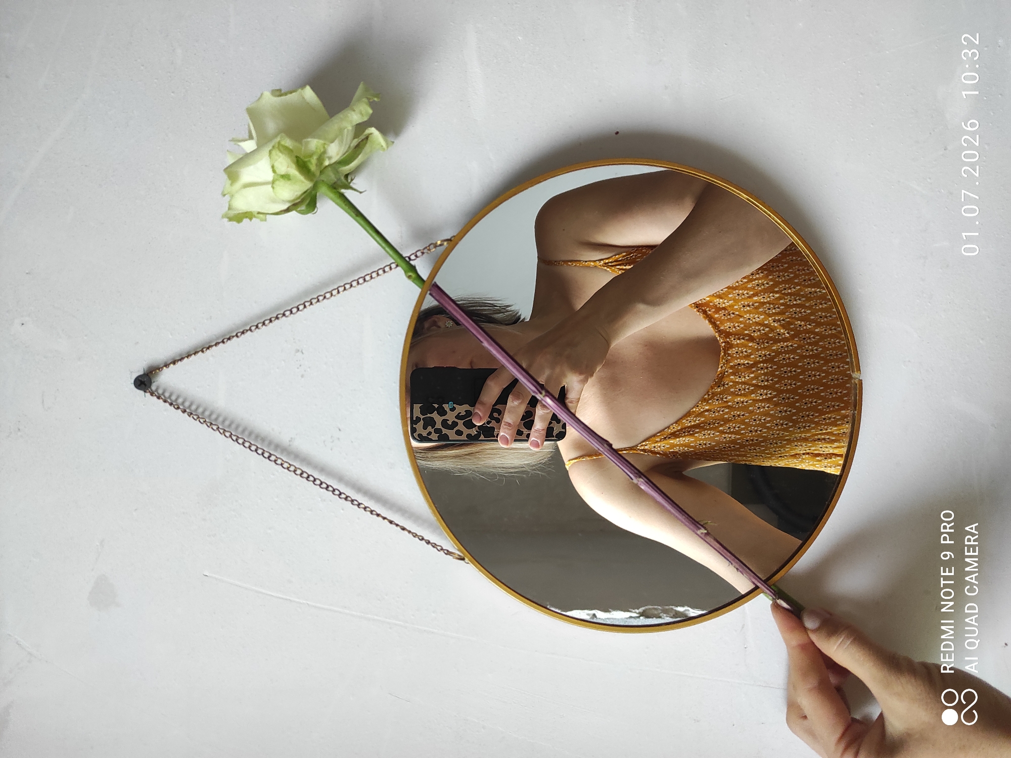 In Malyi Bukor, Russia, a person's torso and arm are visible in the reflection of a round mirror hanging on a textured white wall. The person is holding a pale green rose stem, which is partially obscuring their reflection. The person is wearing a yellow tank top with a white patterned design. A smartphone with a leopard-print case is being held by the person's reflected hand, suggesting they are taking the photograph. The mirror has a thin gold frame and is suspended by a chain. The overall scene is lit by natural light, casting soft shadows.