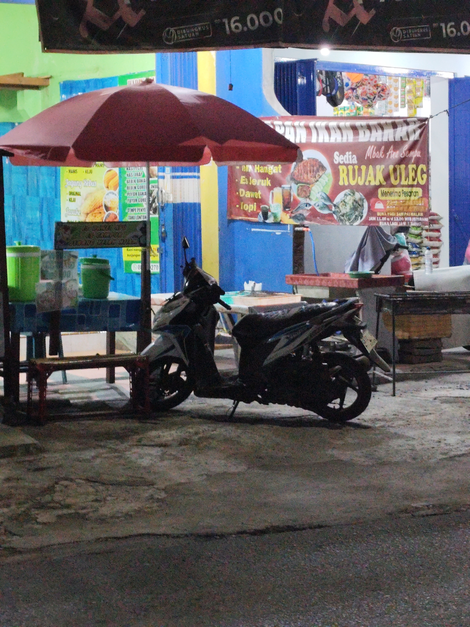 The image depicts an outdoor scene at night, likely a food stall, in Sempu, Indonesia. A dark-colored motorcycle is parked in the foreground, angled to the right. Behind the motorcycle, a stall is set up with a red umbrella providing cover. A table with various items, including green containers and possibly food, is visible under the umbrella. In the background, there is a building with blue and yellow accents and signage that is partially obscured. One sign prominently displays "RUJAK ULEG" and mentions "Sedia Mbak Ana Sempu," suggesting it is a Rujak Uleg stall run by someone named Ana in Sempu. The ground appears to be asphalt or concrete. The lighting suggests artificial illumination, typical of a nighttime setting.
