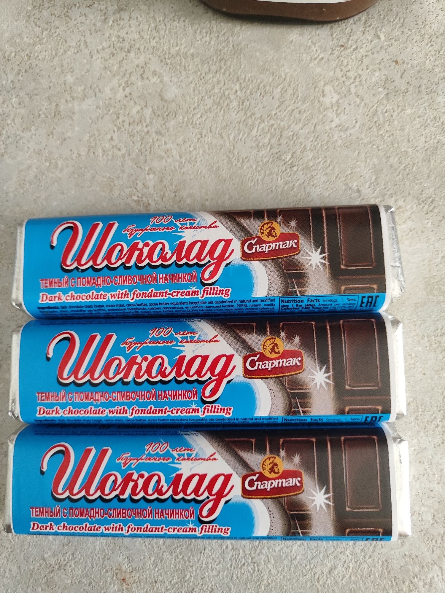 Three chocolate bars are stacked vertically on a light-colored textured surface, possibly a countertop or floor. The chocolate bars are wrapped in blue and silver foil with red lettering. The top bar's wrapper is slightly creased and torn. The brand name "Шоколад" (Shokolad) is prominently displayed in large red cursive font on each wrapper, followed by "Спармak" (Spartak) in a smaller font. Below the brand name, there is text in Russian and English describing the product as "Dark chocolate with fondant-cream filling". The background is out of focus. There are no people or other objects visible in the image.