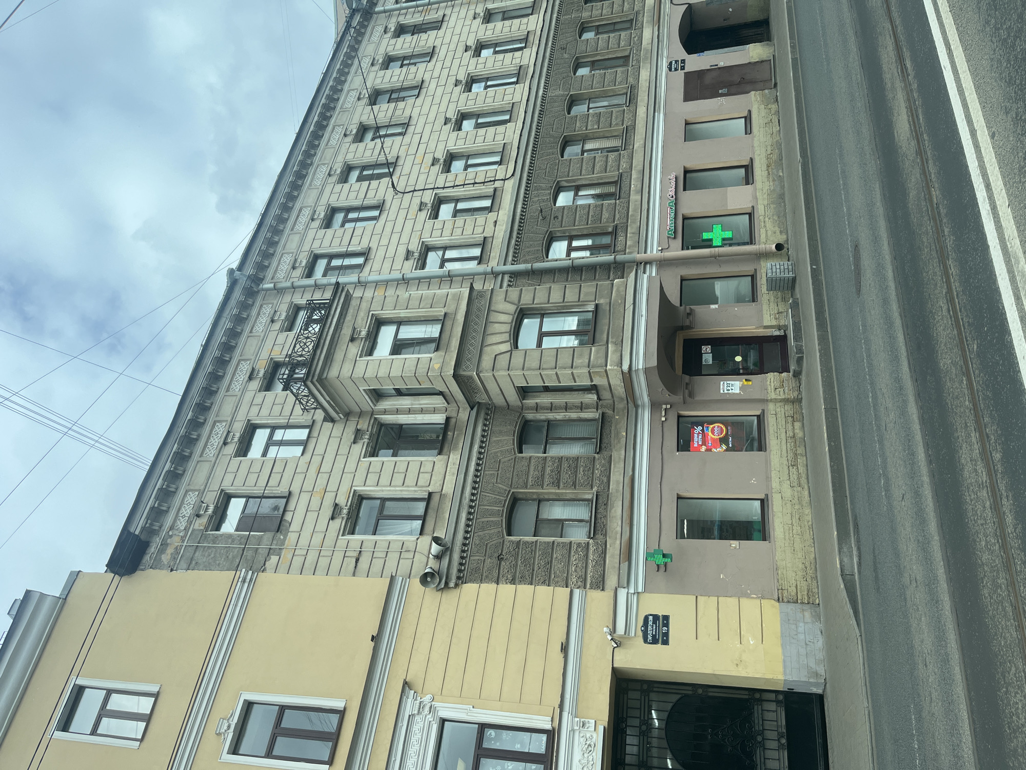 This is a slightly tilted, eye-level, medium shot of a building facade in Saint Petersburg, Russia. The building is tall and appears to be an older architectural style with ornate detailing. It is composed of at least two sections with different facade materials and colors. The left section is a lighter, grayer stone with a grid of regularly spaced windows. The middle section is a darker, textured stone with some windows featuring arched tops. The right section has a smoother, lighter brown facade with a few shops on the ground floor. Visible signs include a bright green cross, indicating a pharmacy, and some advertisements on a window. The building is adjacent to a road with tram tracks. The sky is overcast with clouds, suggesting a cloudy day. The scene is urban, with a focus on the architectural details of the building and its immediate surroundings. There are no people visible. The overall mood is neutral, depicting an everyday urban scene. The visible text includes "Аптека" (Pharmacy) on the green cross sign and "СТАРО-ПЕТЕРГОФСКИЙ" along with a street number "19" on a street sign. There are also some smaller signs with Cyrillic text that are not clearly legible.