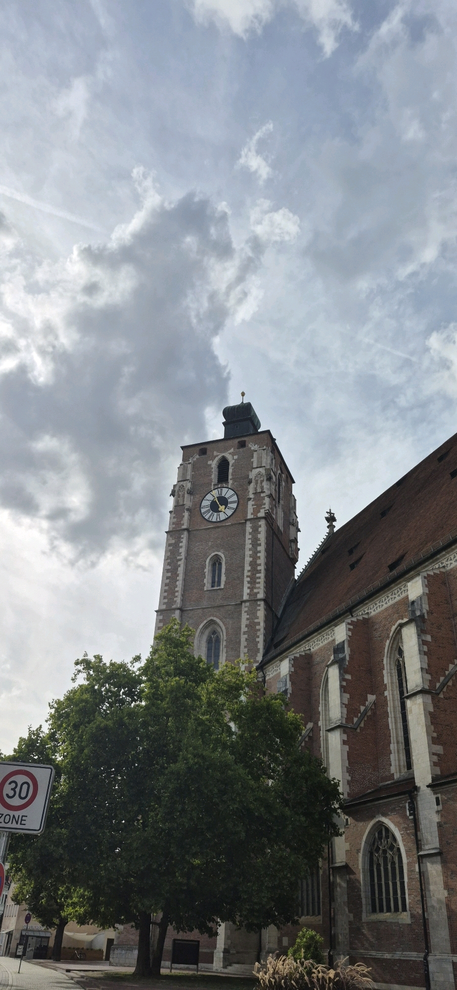The image shows a view of the Neues Rathaus (New Town Hall) in Ingolstadt, Germany. The Neues Rathaus is a large brick building with a prominent clock tower. The clock tower is made of brick and has a pointed roof. The clock face is visible and shows the time as approximately 1:50. In front of the Neues Rathaus is a large tree with green leaves. A traffic sign indicating a "30 ZONE" is visible to the left of the tree. The sky is cloudy.
