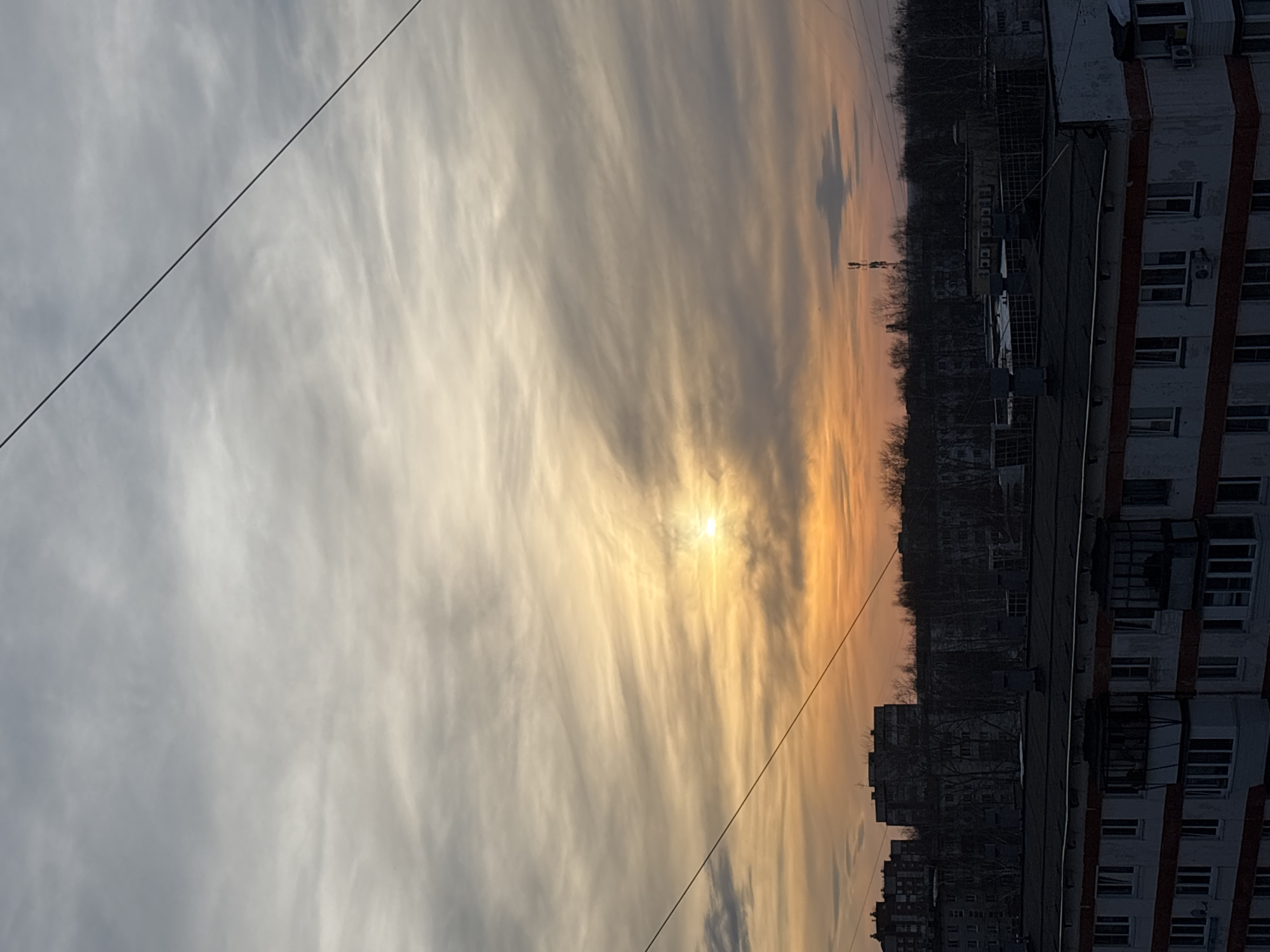 The image presents an urban landscape at sunrise or sunset within the city of Chaikovsky, Russia. The sky displays stratified, wispy clouds, transitioning from grey-white in the upper portion to an intense golden orange hue around a radiant sun, which is visible as a bright disk positioned slightly right of the frame's vertical center. A prominent dark utility cable diagonally traverses the upper sky from the upper-left to the lower-right.
In the foreground, the upper floors and dark, flat roof of a multi-story residential building are visible. This structure features white facades with a distinct horizontal red-orange stripe, alongside multiple dark-framed windows and several balconies or enclosed loggias. The background consists of silhouetted multi-story urban buildings interspersed with a dense line of bare, deciduous trees. A tall telecommunications tower is visible above the tree line in the mid-right background. No human figures or specific actions are depicted.