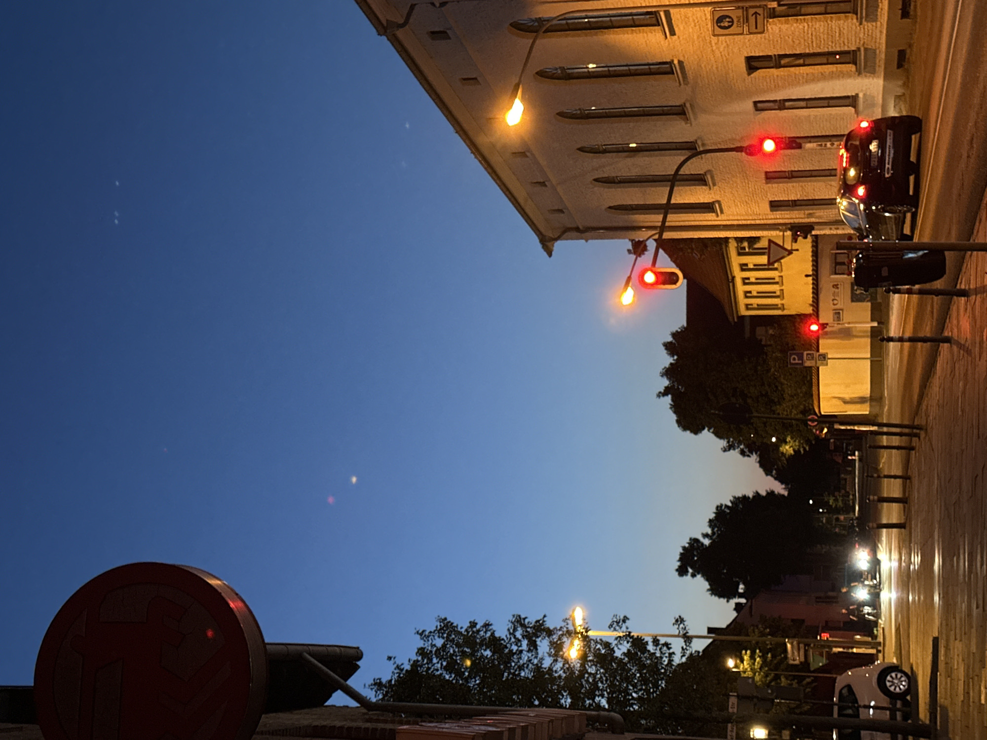 The image shows a street scene in Krefeld, Germany, during twilight. The sky is a deep blue, transitioning to lighter hues near the horizon. A red circular sign with a white graphic is visible in the upper left quadrant, attached to a building. Streetlights illuminate the scene with a warm glow.
In the middle ground, a white building with arched windows stands on the right. Traffic lights are visible, with red lights illuminated. A black car is driving on the road. To the left of the road, a sidewalk is paved with stone. In the background, silhouetted trees and buildings are present.
Further left, a white car is parked on the side of the road. Two figures on what appear to be motorcycles or scooters are in motion, their headlights on. The overall impression is a quiet urban environment at dusk.