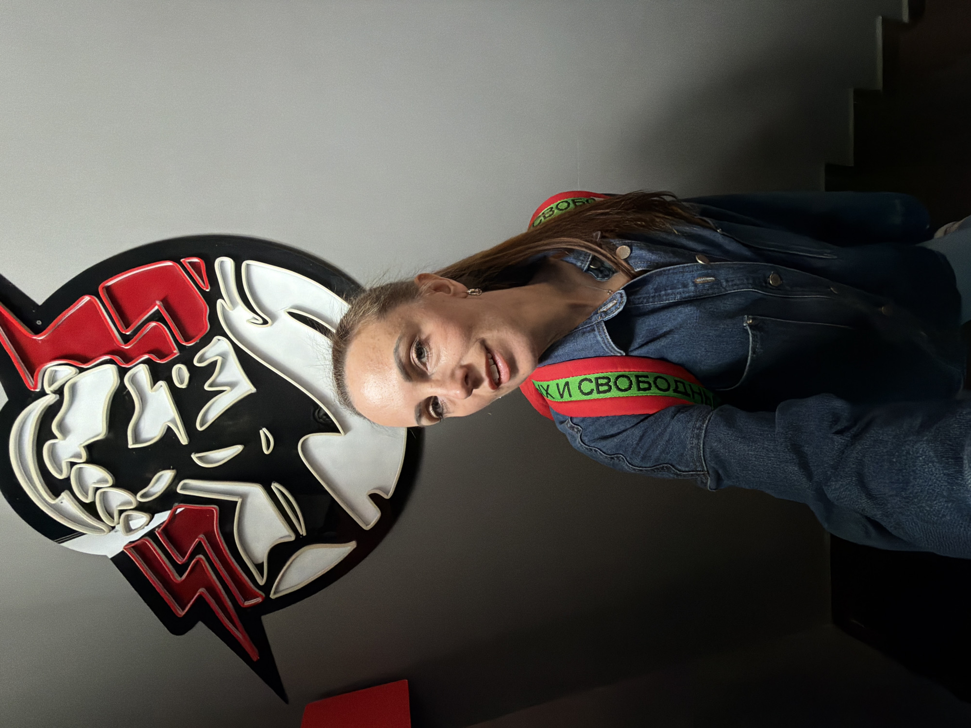 A medium shot shows a woman with her hair in a ponytail, wearing a denim jacket and a red backpack, standing in front of a wall with a large, stylized graphic. The graphic is black, white, and red, with a lightning bolt design. The woman is looking at the camera and smiling. The background is a plain gray wall. The image appears to be taken indoors, possibly in a public space. There is no specific indication that this location is in Birobidzhan, Russia, nor is there any visual information to support this claim.