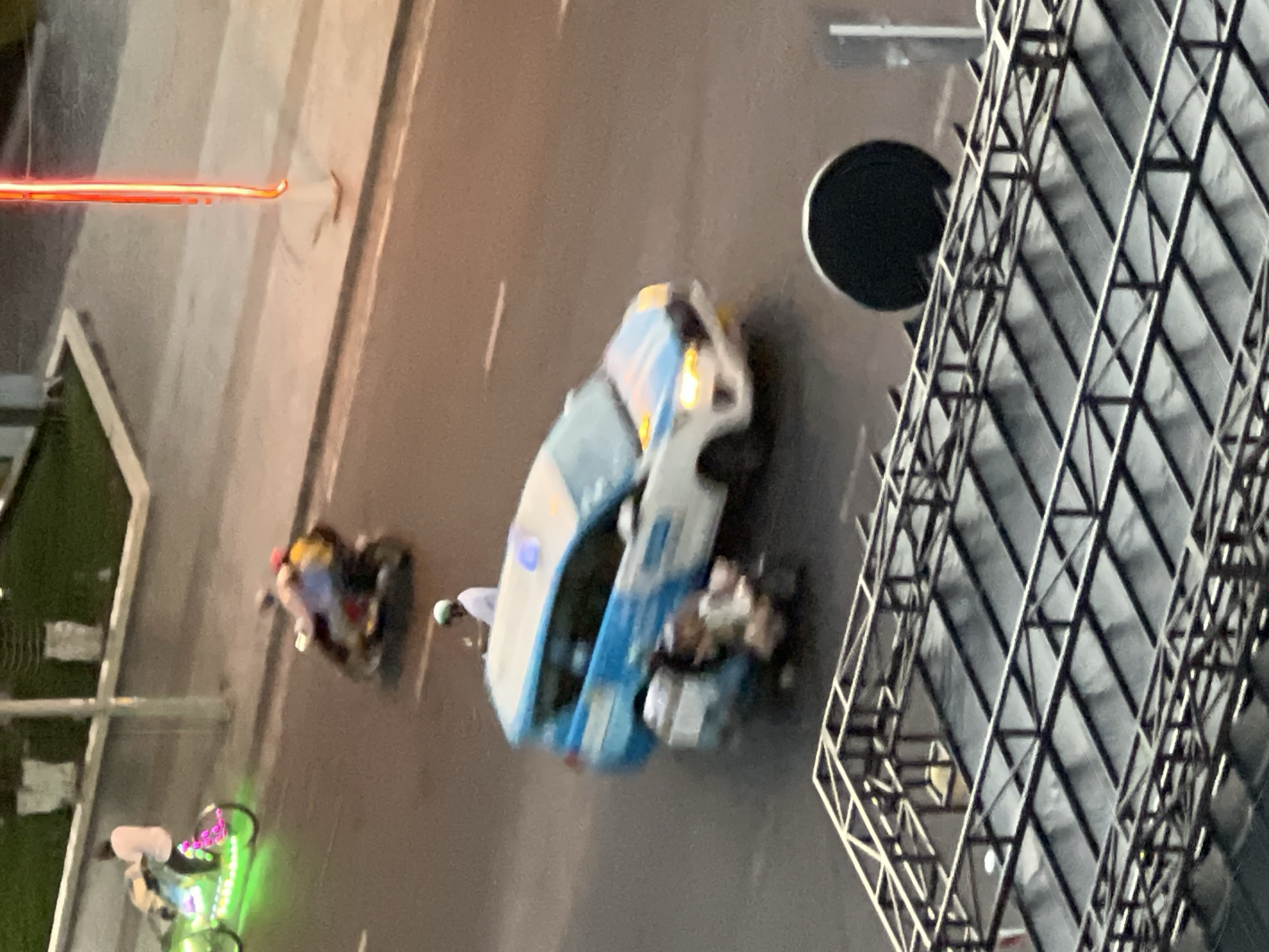 An aerial perspective captures a street scene in Nha Trang, Vietnam. In the foreground, a metal structure, possibly part of a stage or scaffolding, frames the lower edge of the image. Beyond this, the street is visible with a few vehicles and individuals. A blue and white van, possibly a taxi, is the most prominent vehicle, with its headlights illuminated. Two people appear to be on a scooter or motorcycle behind the van, their forms blurred due to motion. Further up the street, another motorcycle with neon green and pink lights is moving, carrying at least two individuals. A red neon light sign is also visible on the right side of the frame. The ground is asphalt, with a sidewalk and some greenery visible in the background. The lighting suggests it is either dawn or dusk.