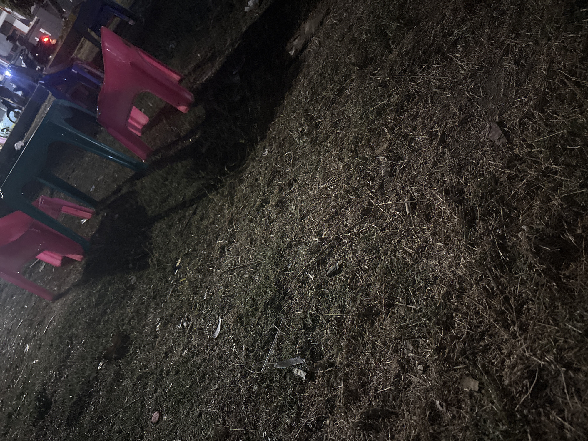 A ground-level shot of a grassy area at night, likely a park or public space in Palimanan, Indonesia. The grass is dry and sparse, littered with debris including small pieces of plastic and leaves. In the background, there are several brightly colored plastic chairs, predominantly pink and blue, arranged around tables. One pink chair is tilted on its side. A teal-colored table is visible, casting shadows. In the far background, there are indications of vehicles, possibly motorcycles, and lights, suggesting activity or a nearby road. The overall scene is dimly lit, with artificial light sources illuminating the objects in the foreground and midground.