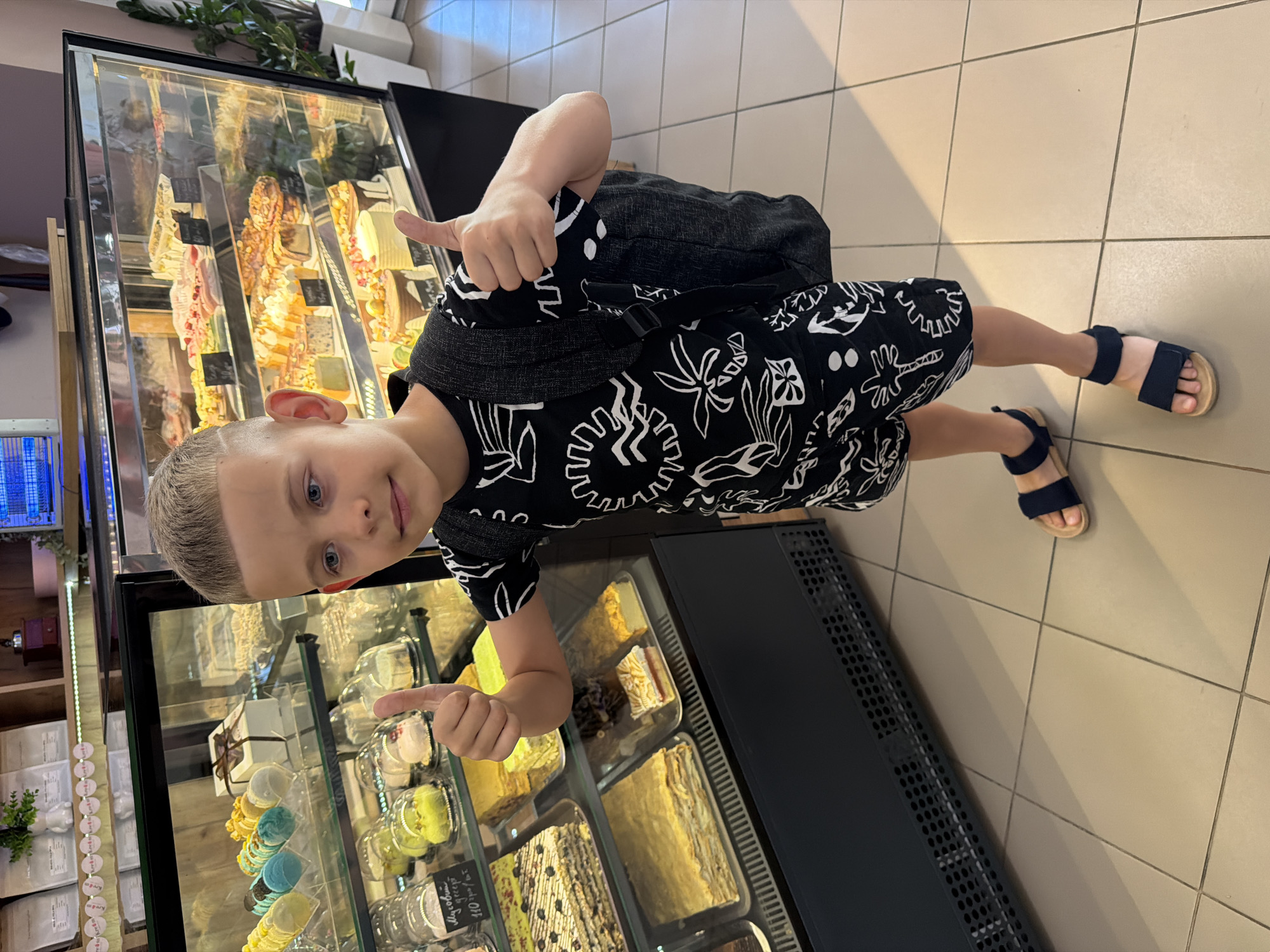 A young boy with blonde hair and blue eyes is standing in a brightly lit indoor space, possibly a bakery or dessert shop, with his body turned towards the camera. He is wearing a black t-shirt with a white floral pattern and dark shorts, and blue sandals. He has a backpack on his back. The boy is smiling and giving a thumbs-up with both hands.
In the background, there are display cases filled with various desserts, including cakes, pastries, and possibly macarons. The display cases are illuminated, showcasing the colorful treats. One display case appears to be a refrigerated unit with a glass front. The floor is tiled with a neutral beige color. There are shelves with stacked items to the left of the boy, and a dark, possibly metal, structure to his right.
The lighting suggests it is daytime. The overall mood of the image is cheerful and positive, conveyed by the boy's smile and gesture. There is no visible text in the image.
