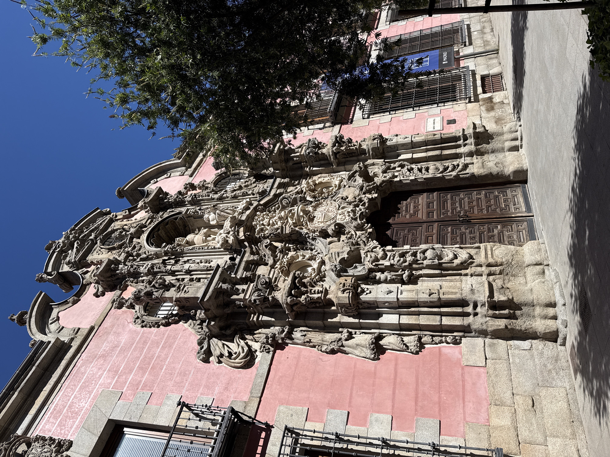 A low-angle shot captures the facade of a pink building in Madrid, Spain, identified by a sign stating "Madrid". The architecture exhibits ornate stone carvings around a large wooden door, with stone detailing extending down the side of the building. Balconies with metal railings are present. The sky is clear and blue, and a tree branches into the top left of the frame, partially obscuring the upper part of the building. The composition is taken from a street-level perspective, including portions of a sidewalk along the right edge of the image.