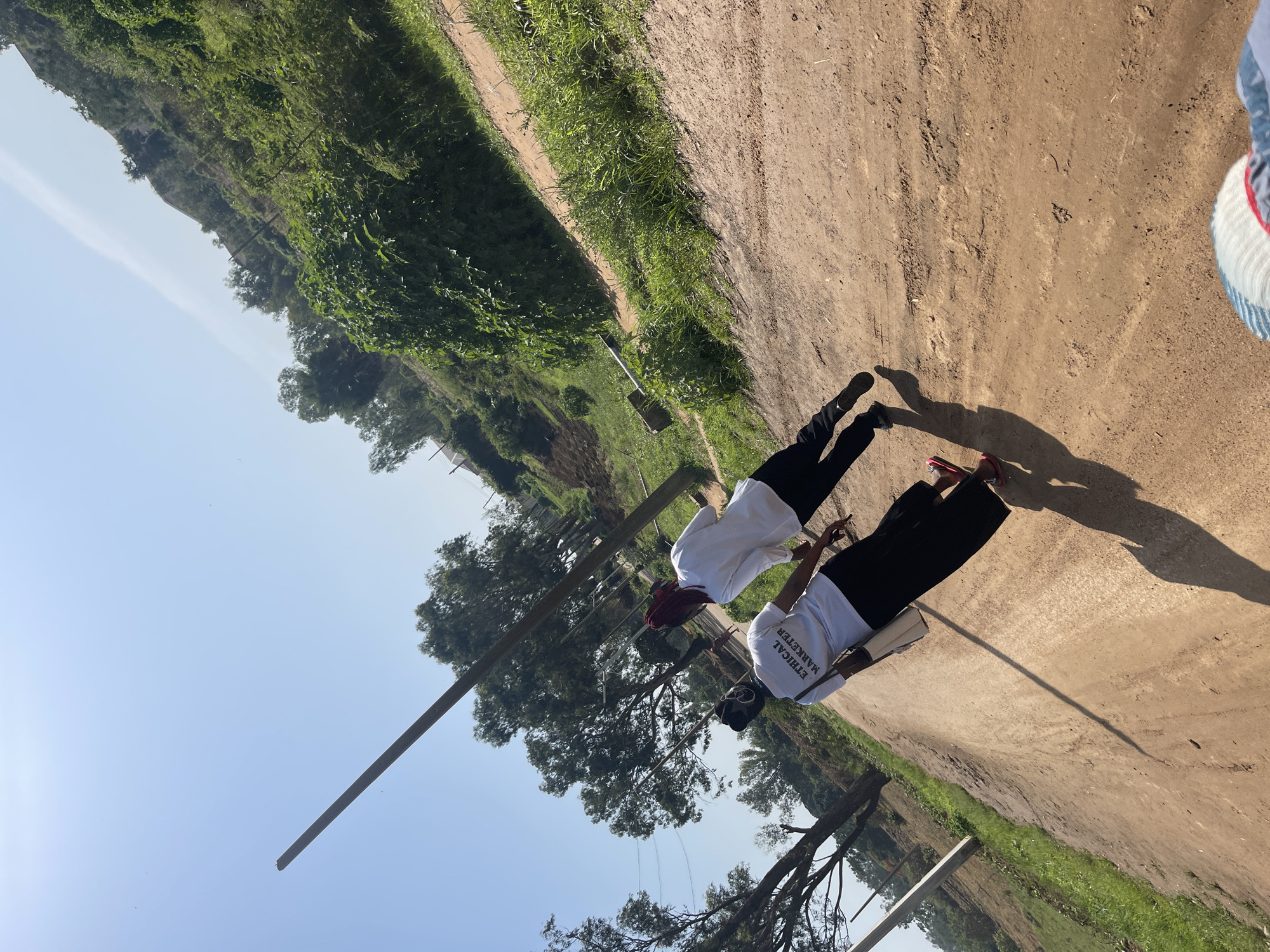 Two individuals are walking away from the camera on a dirt road, which is likely located in the city of Rafin Par, Nigeria. The individual in the foreground is wearing a white t-shirt with the words "ETHICAL MARKETER" printed on the back and dark, wide-legged pants. They are also carrying a light-colored bag over their shoulder. The second individual is wearing a white long-sleeved shirt and black pants. Both individuals appear to be walking in the same direction. The environment is rural, with lush green vegetation on the right side of the road and trees lining the left side. A wooden pole, likely a utility pole, is visible on the left side of the image. The sky is clear and blue, indicating a sunny day. The ground is uneven and shows signs of tire tracks, suggesting it is a frequently used road. The overall scene depicts a casual outdoor walk in a natural setting.
