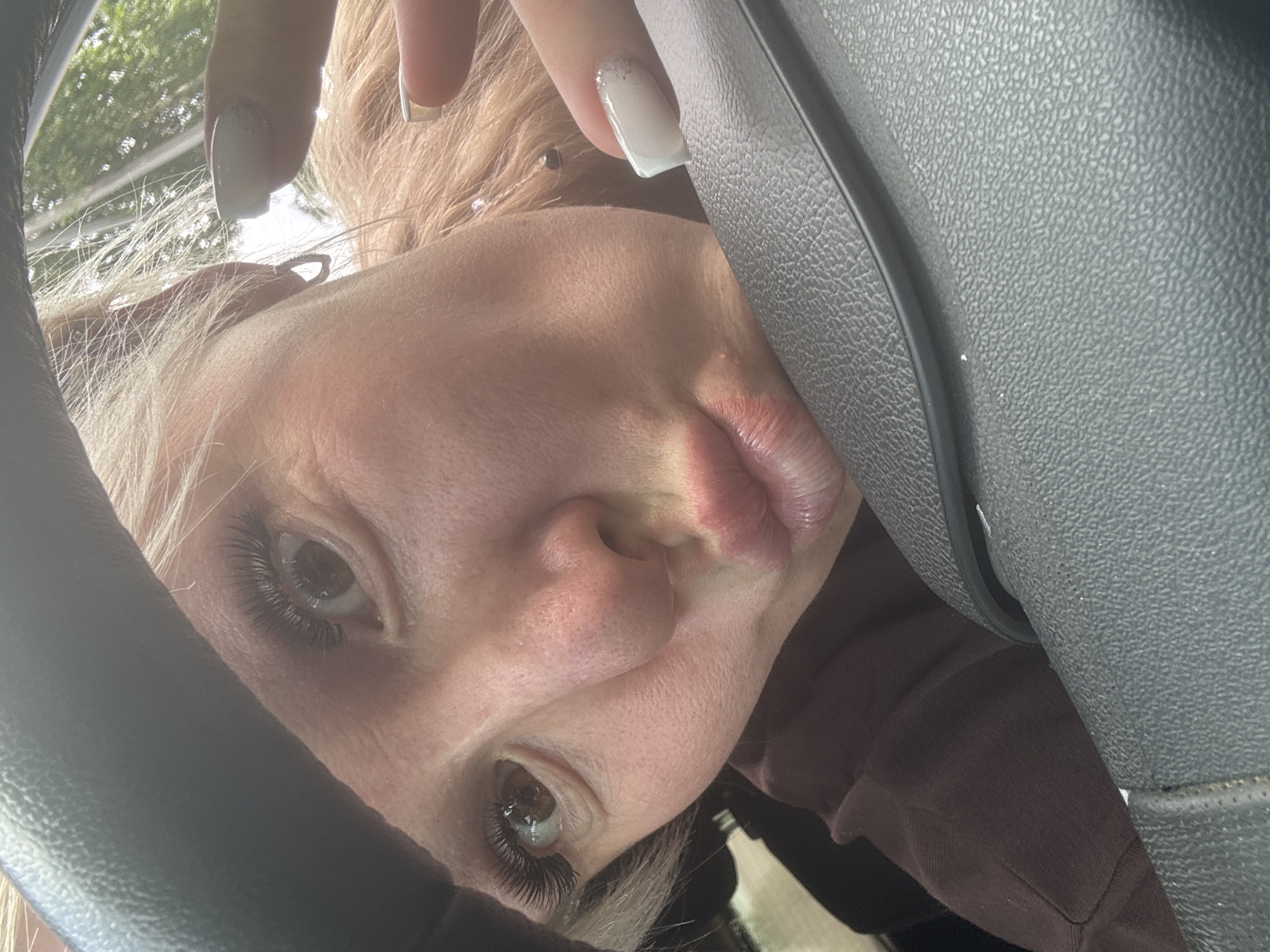A close-up, high-angle selfie shows a young blonde woman in the foreground, positioned in the driver's seat of a car. She is leaning forward, her face angled towards the camera, with her lips puckered as if for a kiss. Her eyes are open, and she has on dramatic false eyelashes. She is wearing a dark brown long-sleeved shirt, and her hair is light blonde and somewhat messy. Her right hand is visible near the top right of the frame, with painted white fingernails, and appears to be touching the steering wheel. The steering wheel is dark grey and textured. The background shows a blurry green outdoor scene, suggesting trees and foliage, possibly indicating a location within the city of Nozhovka, Russia, during daylight hours.