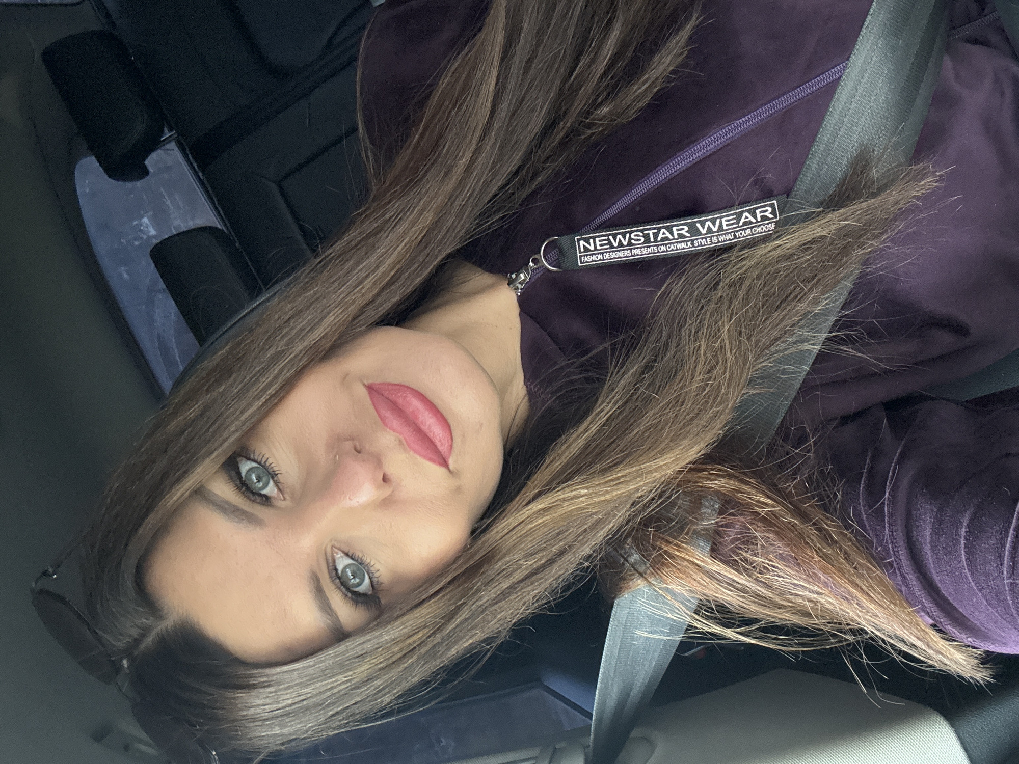 A woman with long brown hair and blue-green eyes is pictured inside a vehicle, wearing a dark purple hooded sweatshirt with a zipper and a gray seatbelt. She has on makeup, including pink lipstick and eyeliner. Sunglasses are resting on top of her head. The interior of the car is visible behind her, with headrests and tinted windows. The woman appears to be in the city of Chaikovsky, Russia, given the context provided.