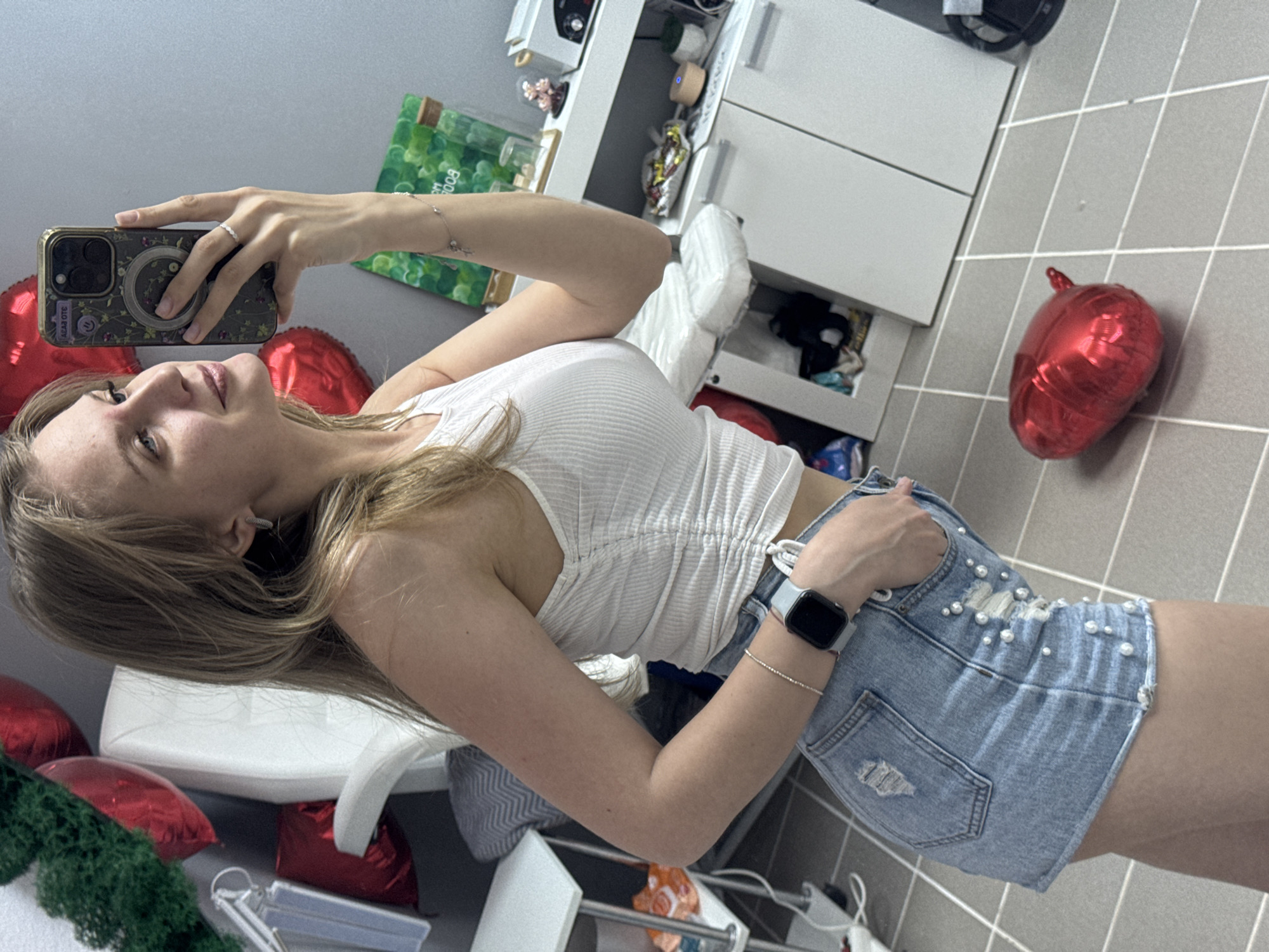 A young woman with blonde hair is taking a selfie with her phone. She is wearing a white top and denim shorts with pearl embellishments. There are several red heart-shaped balloons and some green foliage in the background. A white dresser with drawers is visible in the background. The location is difficult to determine due to the limited visual information. There are no identifying markers to suggest Izhevsk, Russia. The photograph appears to be taken indoors.