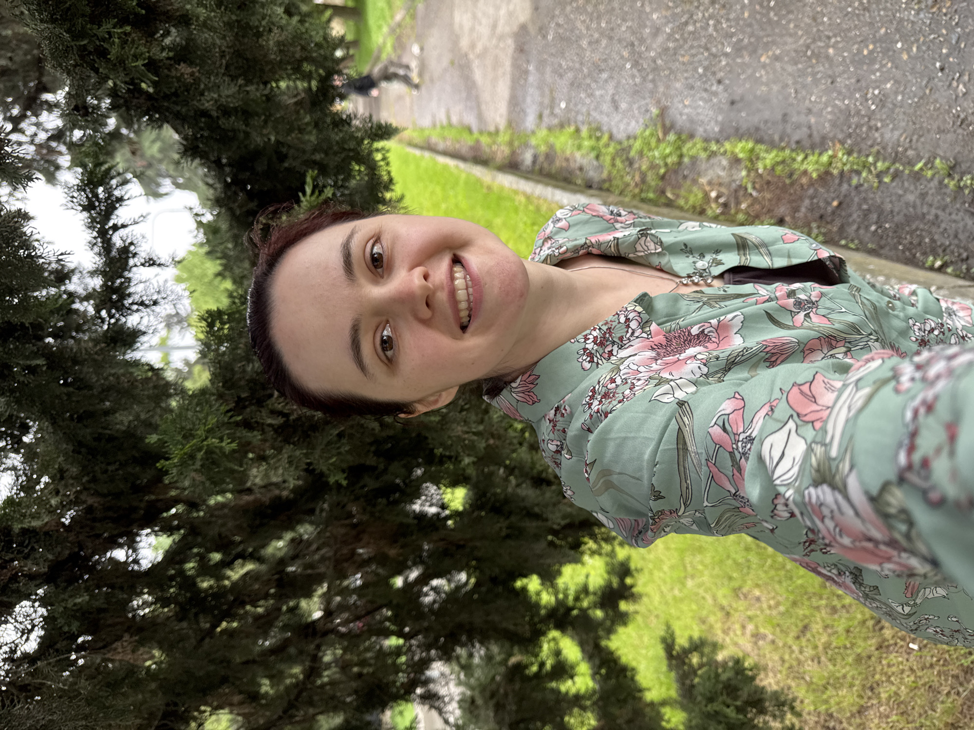 A woman with fair skin and reddish-brown hair pulled back in a bun smiles directly at the camera in an outdoor selfie. She wears a green floral-patterned blouse with pink and white flowers. To her left, dense green trees frame the image. Behind her and to the right, there is a grassy area adjacent to a gray concrete pathway. In the distance along the pathway, there is a person walking away from the camera. The overall impression is a casual, natural setting, potentially within a park or garden. Based on location metadata, this image was taken in Sochi, Russia.