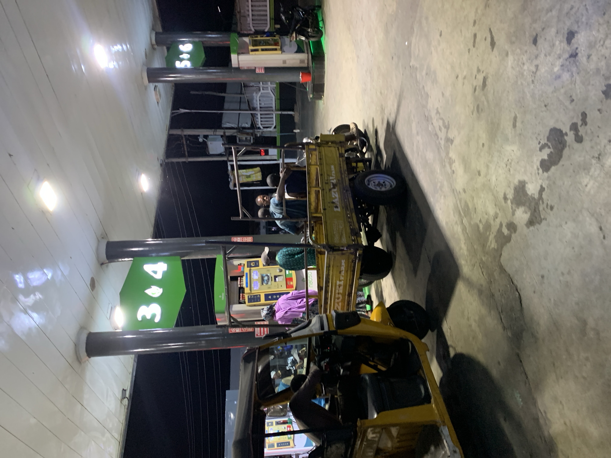 The scene depicts a gas station in Lokoja, Nigeria, at night. The gas station has pumps labeled "3&4" and "5&6". Two three-wheeled auto-rickshaws (tuk-tuks) are parked near the pumps. Several people are visible, including individuals in the back of one tuk-tuk and a driver in the front of the other. The ground is concrete.