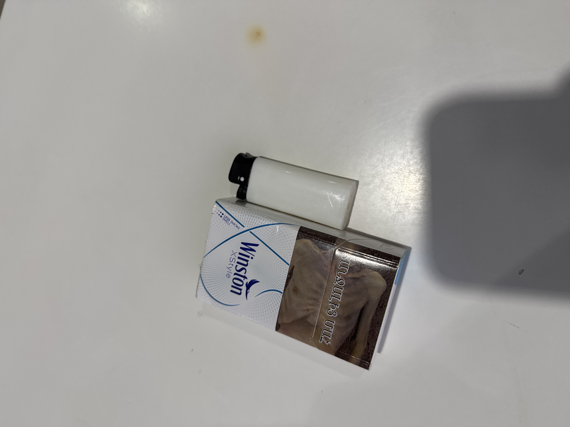 The image displays a pack of "Winston XStyle" cigarettes and a white disposable lighter resting on a plain, light-colored surface, likely a table or desk. The cigarette pack is predominantly white and blue, featuring the brand name "Winston XStyle" and the text "LESS SMOKE SMELL". A graphic health warning is visible on the right side of the pack, depicting an emaciated or skeletal torso, accompanied by Armenian text that reads "ԱՌՈՂՋՈՒԹՅԱՆ ՀԱՄԱՐ" (meaning "FOR HEALTH"). The white lighter, with a black top, is placed directly on top of the cigarette pack.
The scene is indoors, brightly lit, with a noticeable shadow cast on the right side of the frame, suggesting an overhead light source or an object out of frame. The surface appears to be slightly glossy and shows minor imperfections and a small, light brown stain in the upper center. No activity is taking place; the items are stationary. The presence of Armenian text on the cigarette pack strongly indicates the location context of Armenia, specifically Yerevan as provided.
