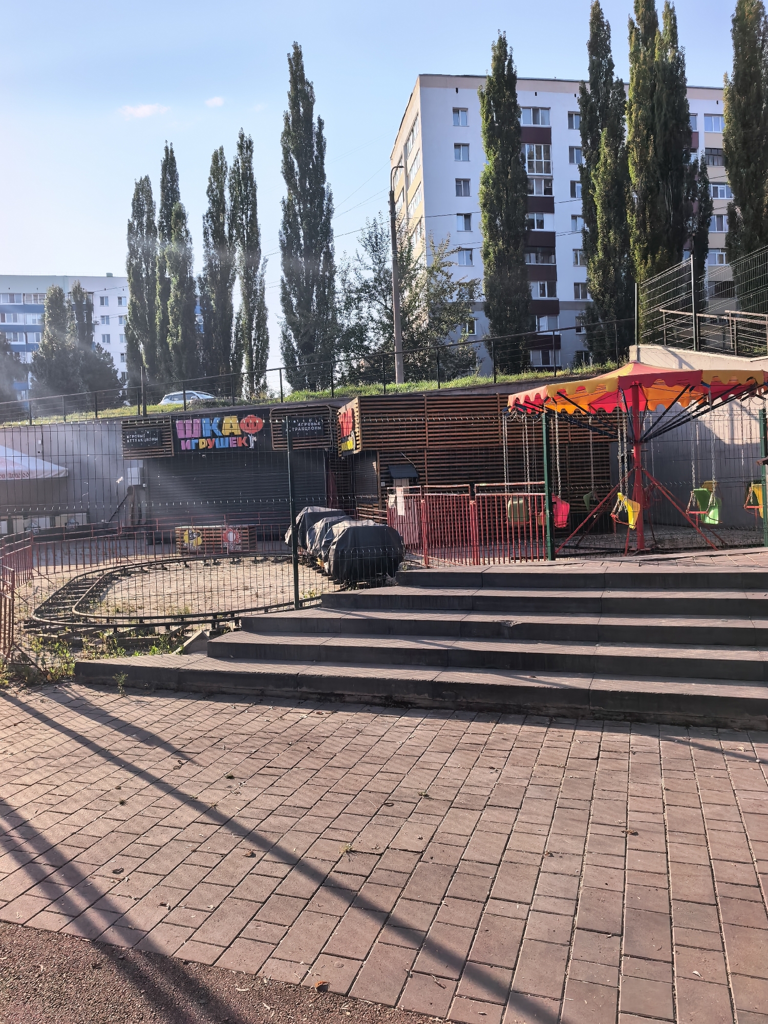 The image shows a playground in Sterlitamak, Russia. The playground features a small train track, a carousel with colorful swings, and a building with signage that reads "ШКАФ ИГРУШЕК" (Shkaf Igrushek), meaning "Toy Cabinet" in Russian. The building also has text that appears to indicate "ИГРОВЫЕ АТТРАКЦИОНЫ" (Igrovye Attraktsiony), meaning "Amusement Rides". Tall trees are visible behind the playground, and apartment buildings are in the background. The ground is paved with bricks in the foreground, and a set of concrete stairs leads up to the playground area. The scene is illuminated by sunlight, casting shadows on the paved ground.