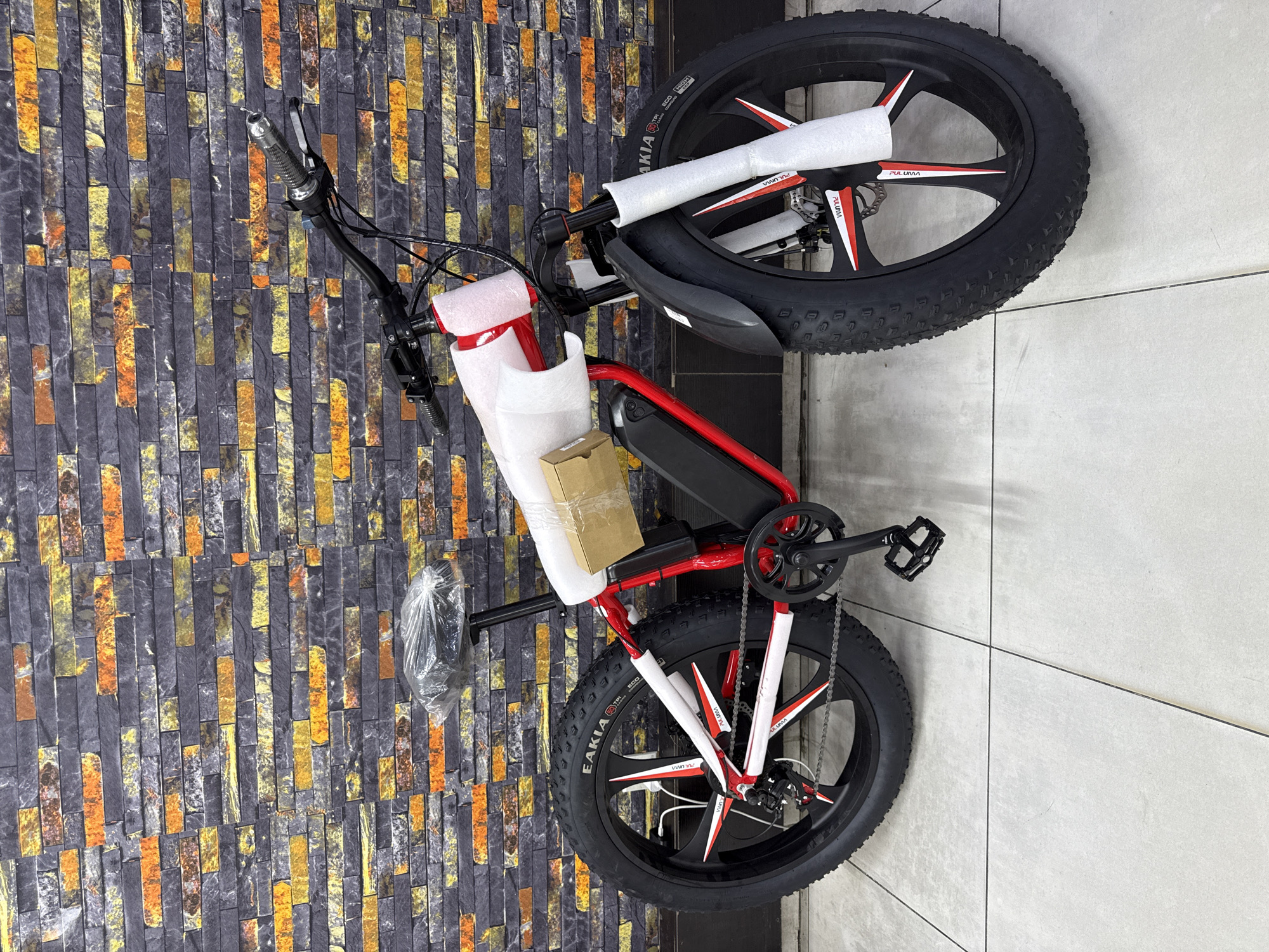 The image depicts a red electric bicycle with black tires, partially wrapped in white protective foam, leaning against a wall with a stone tile design. The environment suggests an indoor setting, possibly a shop or storage area in Ado, Nigeria. A cardboard box is secured to the frame, and the bicycle's wheels feature white and red accents. The background includes a smooth tiled wall, contrasting with the textured wall. No people are visible. The focus is on the bicycle's features and presentation.