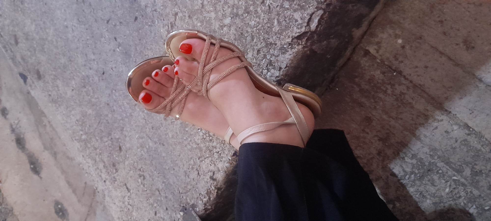 The image displays a close-up view of a person's feet, clad in light gold or champagne-colored flat sandals with multiple glitter-embellished straps across the instep and around the ankle. The person's toes feature a vibrant red nail polish. A section of dark fabric, likely from pants or a skirt, is visible around the lower leg/ankle area.
The feet are resting on an uneven, textured ground surface that appears to be concrete or an old paved area, characterized by varying shades of gray, dark patches, and rough elements. To the right of the feet, a darker, rough-textured, possibly stone or concrete, surface suggests a curb, wall base, or a different ground section. The lighting is somewhat dim, creating subtle shadows, indicating it could be overcast, early evening, or an indoor setting with low light. No specific activity is depicted other than the static pose of the feet. There is no visible text in the image. The scene is set in Juan Griego, Venezuela.