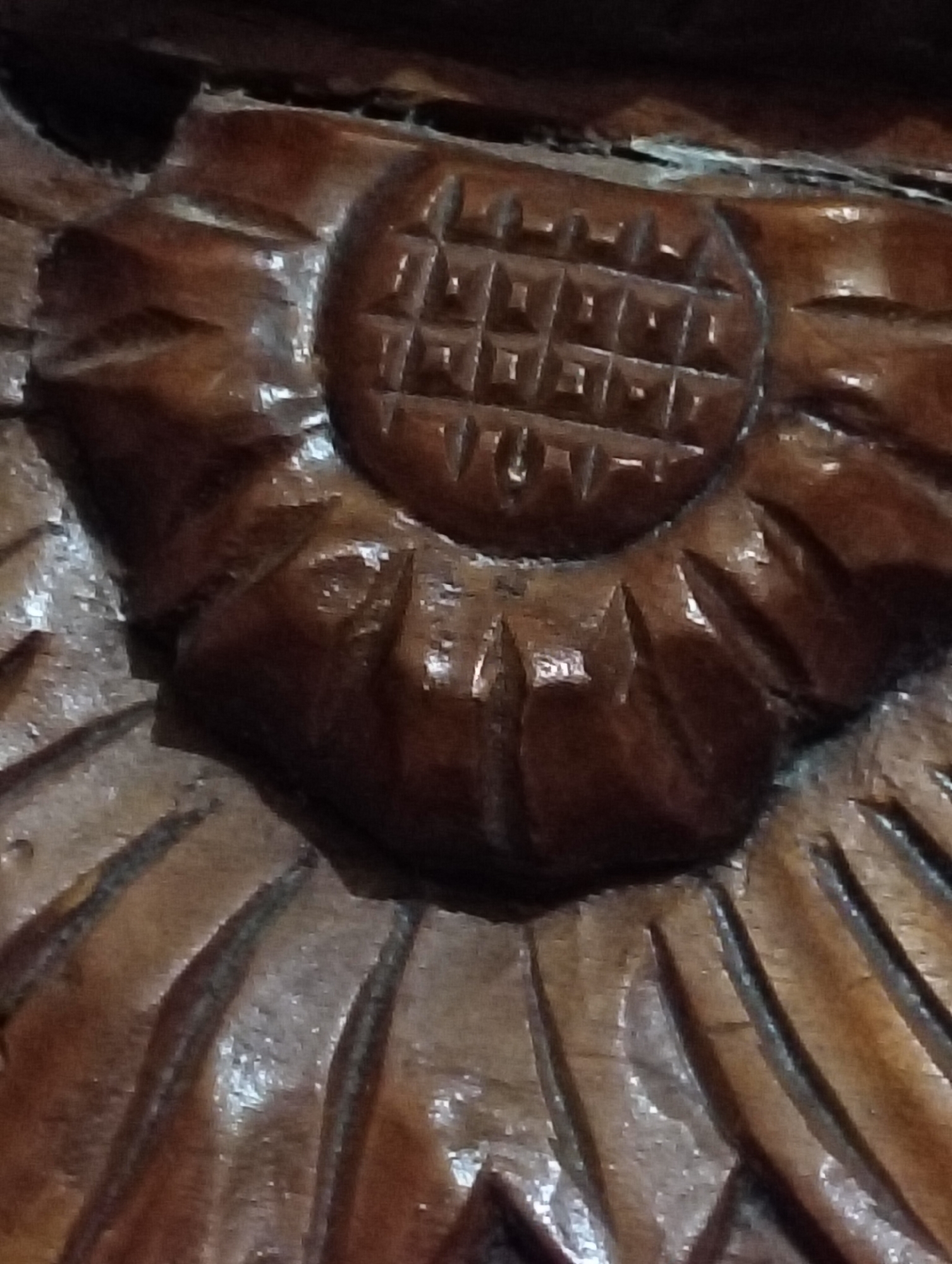 The image shows a close-up of a decorative wooden carving. The carving appears to be of a sunflower or a similar floral design. The petals are deeply incised, and the center of the flower is carved with a grid pattern, suggesting a stylized representation of seeds. The wood is dark brown and shows signs of age and wear, with some areas appearing smoother than others due to polishing or use. Light reflects off the carved surfaces, highlighting the texture and depth of the design. The overall impression is one of intricate craftsmanship. The location in the city of Payagaboh, Indonesia, is not discernible from the image itself; this information is external to the visual content.