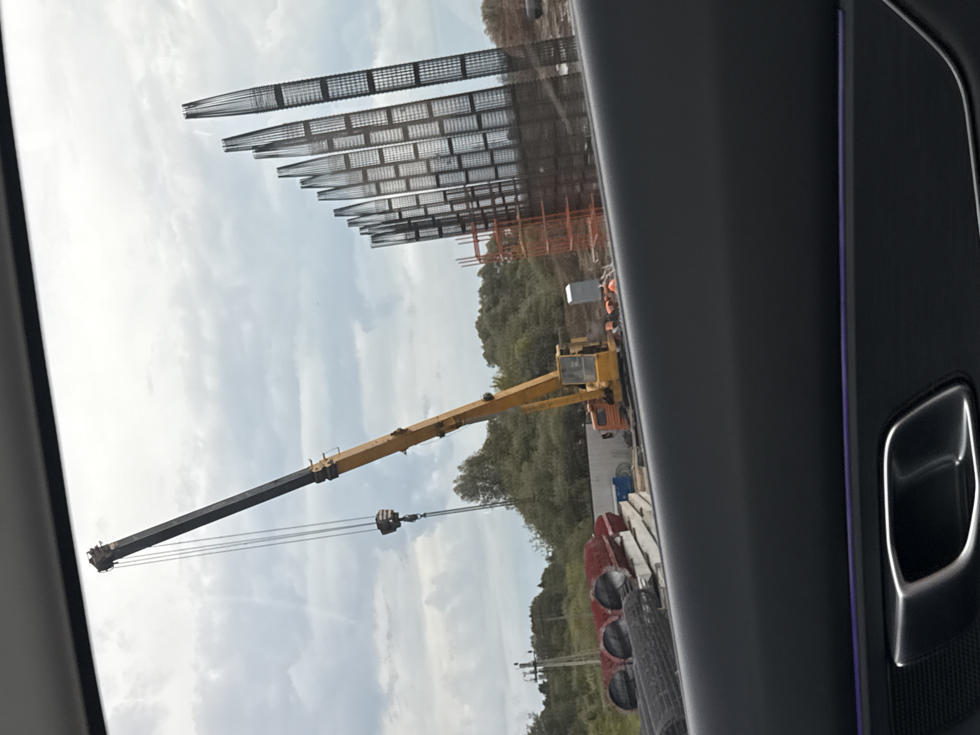 The image captures a construction or industrial site in Kabanikha, Russia, viewed through the window of a vehicle, indicated by the car's dark interior door panel, chrome handle, and a purple ambient light strip visible on the right.
In the background, under a partly cloudy sky, are several large, skeletal industrial structures. These appear to be frameworks, possibly for a large building or facility, composed of numerous vertical and horizontal grid-like sections. To the right of these structures, further construction elements, including some scaffolding, are visible.
In the midground, a prominent yellow and black mobile crane with an extended boom is positioned on the ground, indicating ongoing heavy lifting operations. The crane's hook and cables hang downwards. Scattered around the base of the crane and extending into the foreground are various construction materials, including large red cylindrical objects (possibly pipes or storage containers) and stacks of rectangular, precast concrete or stone blocks, along with rolls of mesh fencing or reinforcement.
Lush green trees and foliage are visible behind the crane and some of the construction materials, suggesting the site is on the edge of a natural area. The time of day appears to be daytime, with soft, diffused light indicating an overcast or partly cloudy sky. No discernible text is visible anywhere in the image. The overall scene depicts an active industrial development or construction project.
