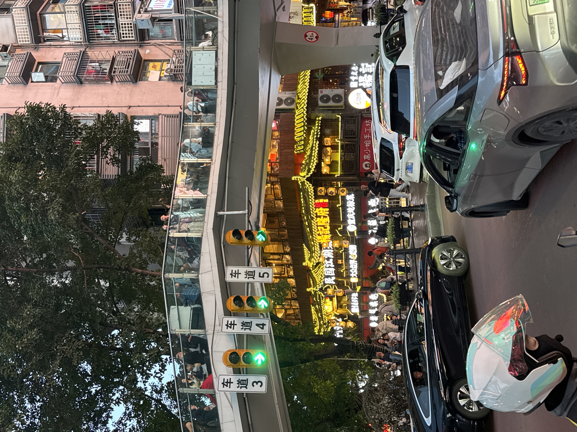 The image depicts a street scene in the Yuzhong District of Chongqing, China. It shows a mix of old and new architecture, with traditional Chinese-style buildings adorned with bright neon lights alongside modern structures. Several vehicles are visible on the street, including cars and a scooter, with people dining outdoors at restaurants, contributing to a lively atmosphere. Above the street scene is an enclosed pedestrian bridge with people using it, adding a layer of complexity to the urban landscape. Traffic lights are on the side of the overpass. The signs indicate that the overpass has 5 lanes. Signs written in Chinese appear on the buildings, indicating local businesses.