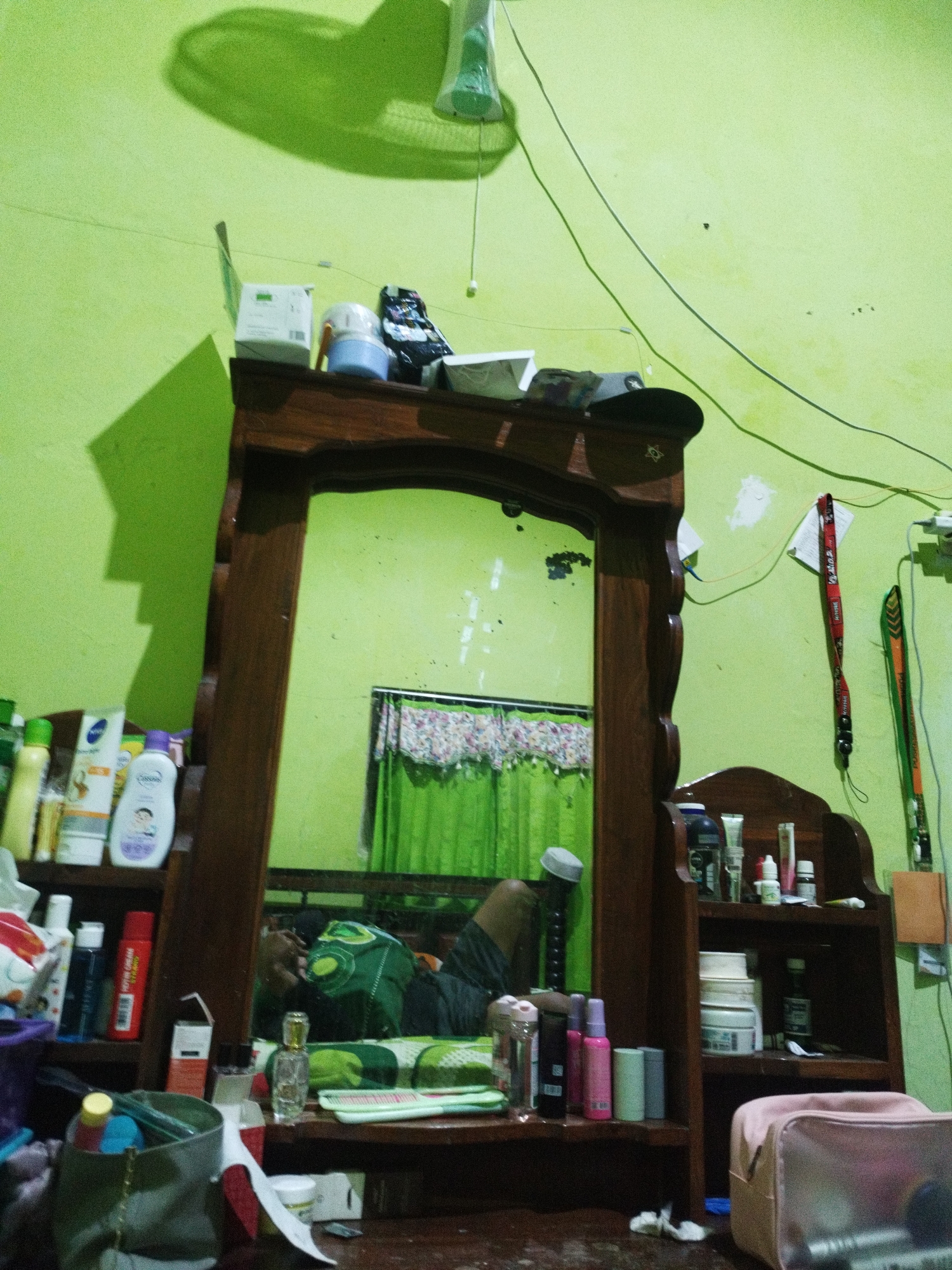 The image depicts a dresser with a mirror, positioned in a room with light green walls in Marengan Daya, Indonesia. The dresser is made of dark wood and holds various cosmetic items, toiletries, and accessories. On the shelves are numerous bottles and containers of skincare products, including lotions, creams, and sprays, some of which are identifiable by their labels such as "Nivea" and "Cussons". A glass perfume bottle is also visible.
The mirror reflects the room, showing a person lying on a bed, partially obscured by a green patterned blanket. A white cap rests on the person's head. Behind the dresser, green curtains with a floral pattern are visible. Above the dresser, a shadow of a ceiling fan is cast on the wall. Electrical wires are strung across the wall. On the right side of the dresser, there is a small wooden shelf unit with more containers. A pink cosmetic bag sits on the dresser's surface in the foreground. The overall scene suggests a personal space, likely a bedroom, where grooming and personal care items are organized.