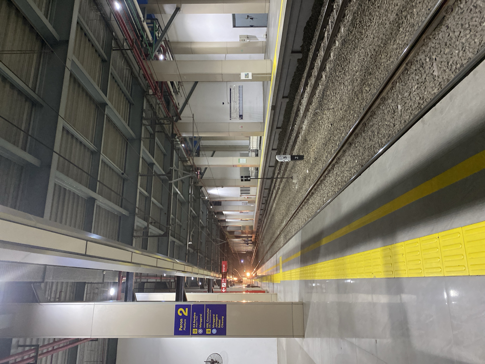 The image depicts an indoor train station platform in Central Jakarta, Indonesia. The platform is marked with yellow tactile paving and a yellow line along its edge. The tracks are visible, with gravel ballast between them. Overhead, a metal roof structure is supported by beige columns. Signs are mounted on a pillar, indicating "Peron 2 Platform" and listing train destinations such as "KA Bandara Airport Train" and "KRL Commuter." A train with illuminated headlights is approaching the platform in the distance. There are no people clearly visible on the platform or tracks, except for a faint, silhouetted figure standing near the approaching train. The environment is well-lit by artificial lights.